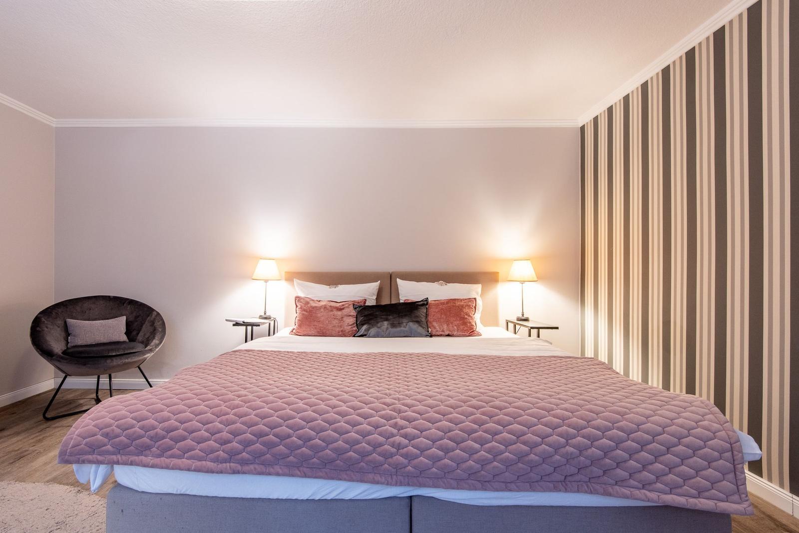 Bedroom with bed, nightstands, and lamps. Striped wall. Chair in corner.