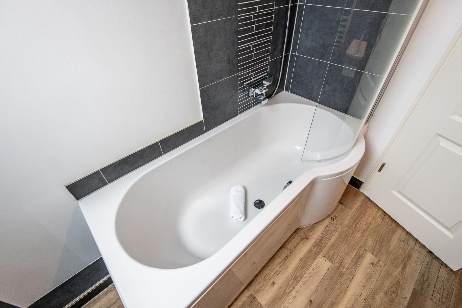 Badewanne mit Duschkabine, dunkle Fliesen und Holzboden.