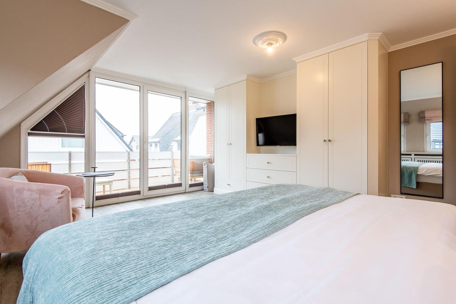 Schlafzimmer mit Bett, Schrank und TV. Große Fenster mit Blick auf Balkon.