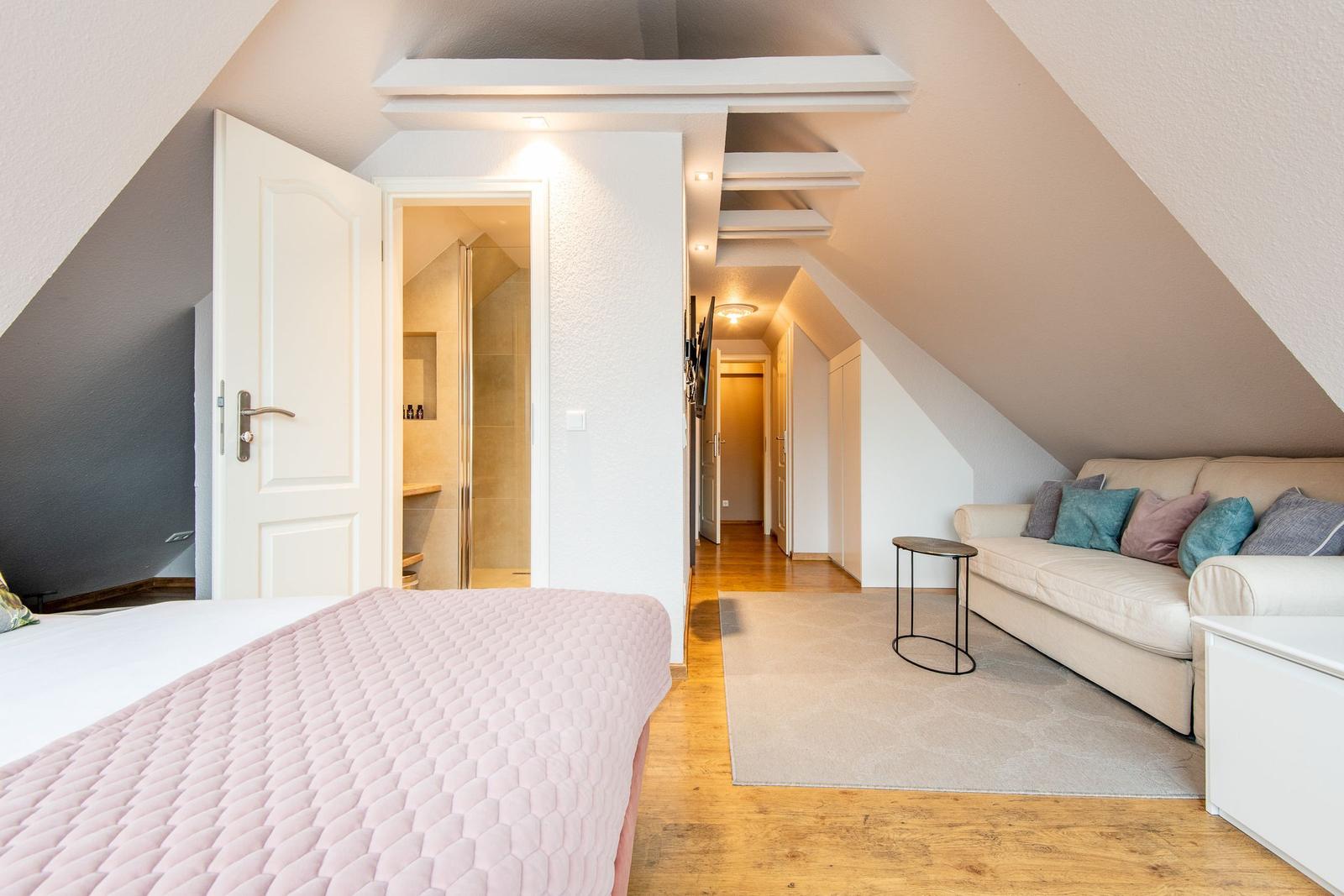 Schlafzimmer mit Bett und Sofa unter Dachschrägen. Blick in Badezimmer und Flur.