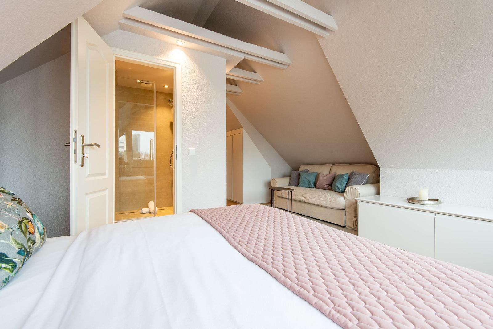 Schlafzimmer mit Dachschrägen, Bett, Sofa und Bad mit Dusche.