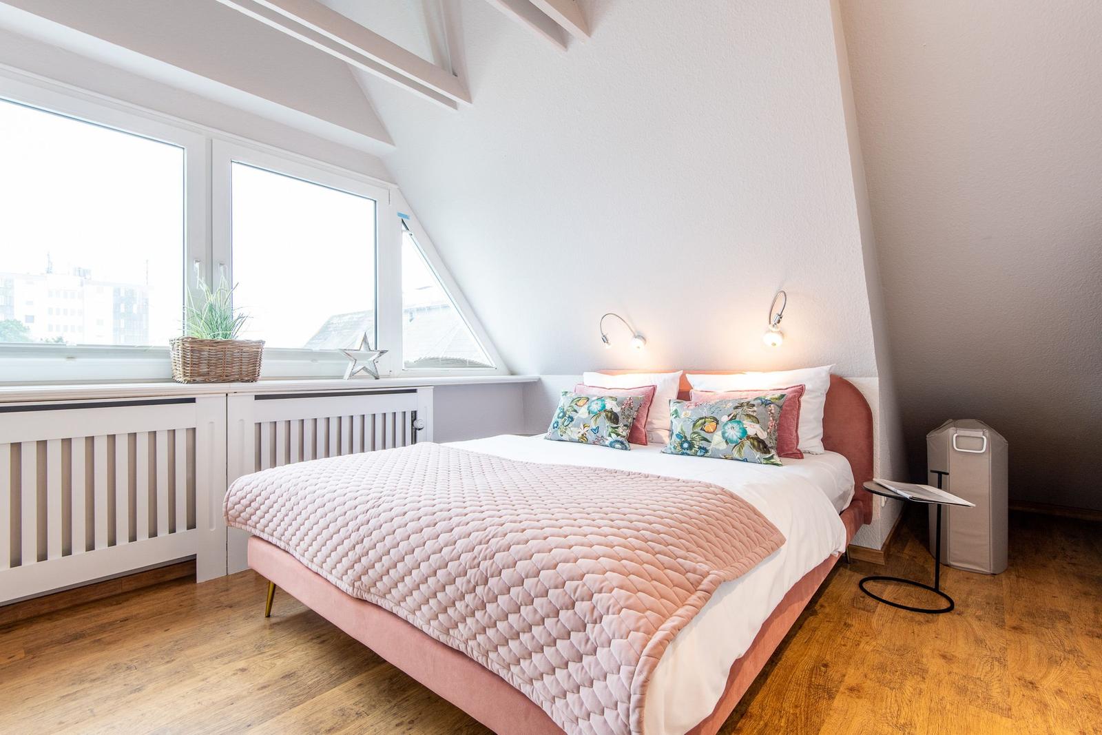 Schlafzimmer mit Dachschrägen, großem Fenster und Bett mit rosa Bezügen.