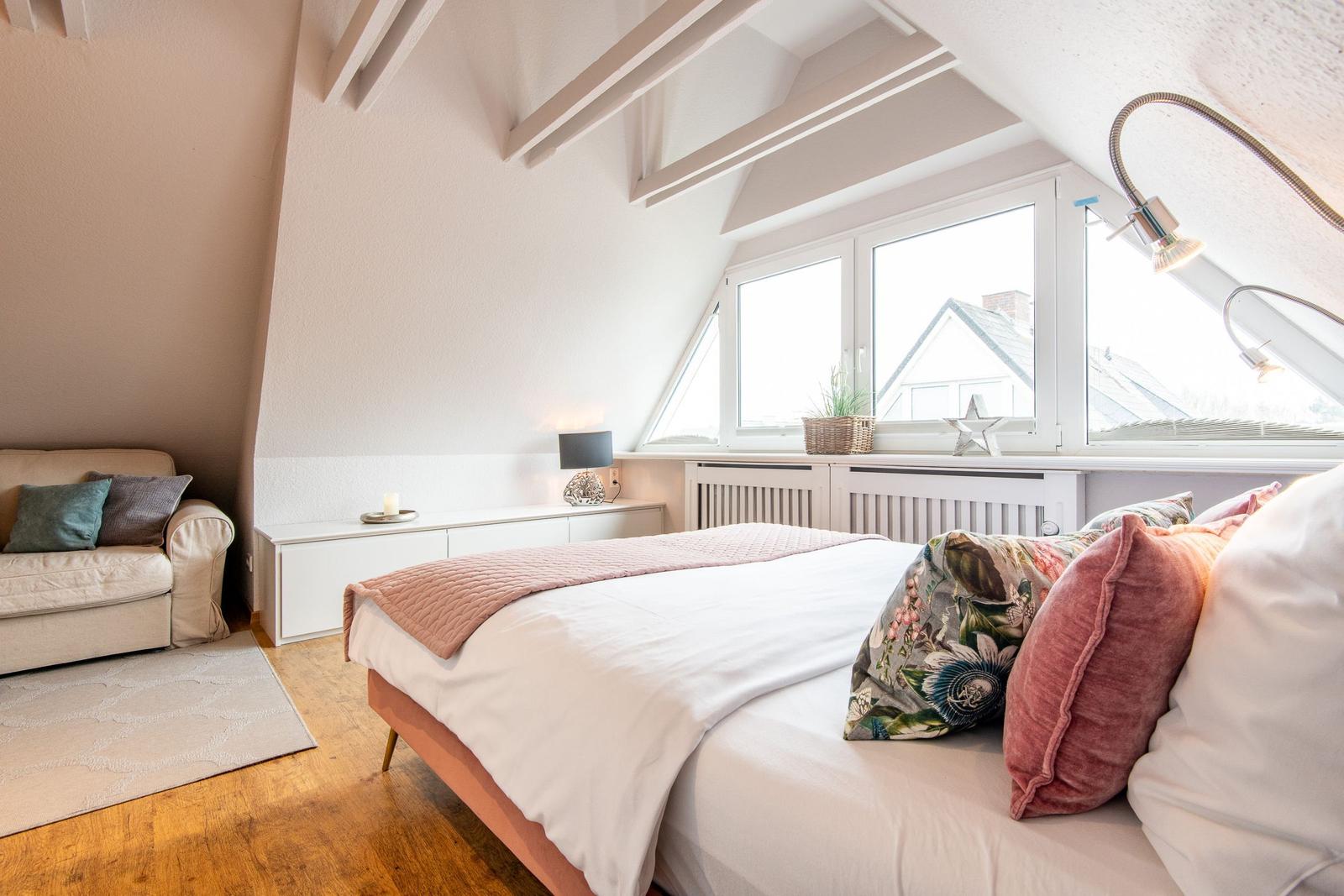 Schlafzimmer mit Dachschrägen, Bett, Sofa und Fenster mit Blick auf Dächer.