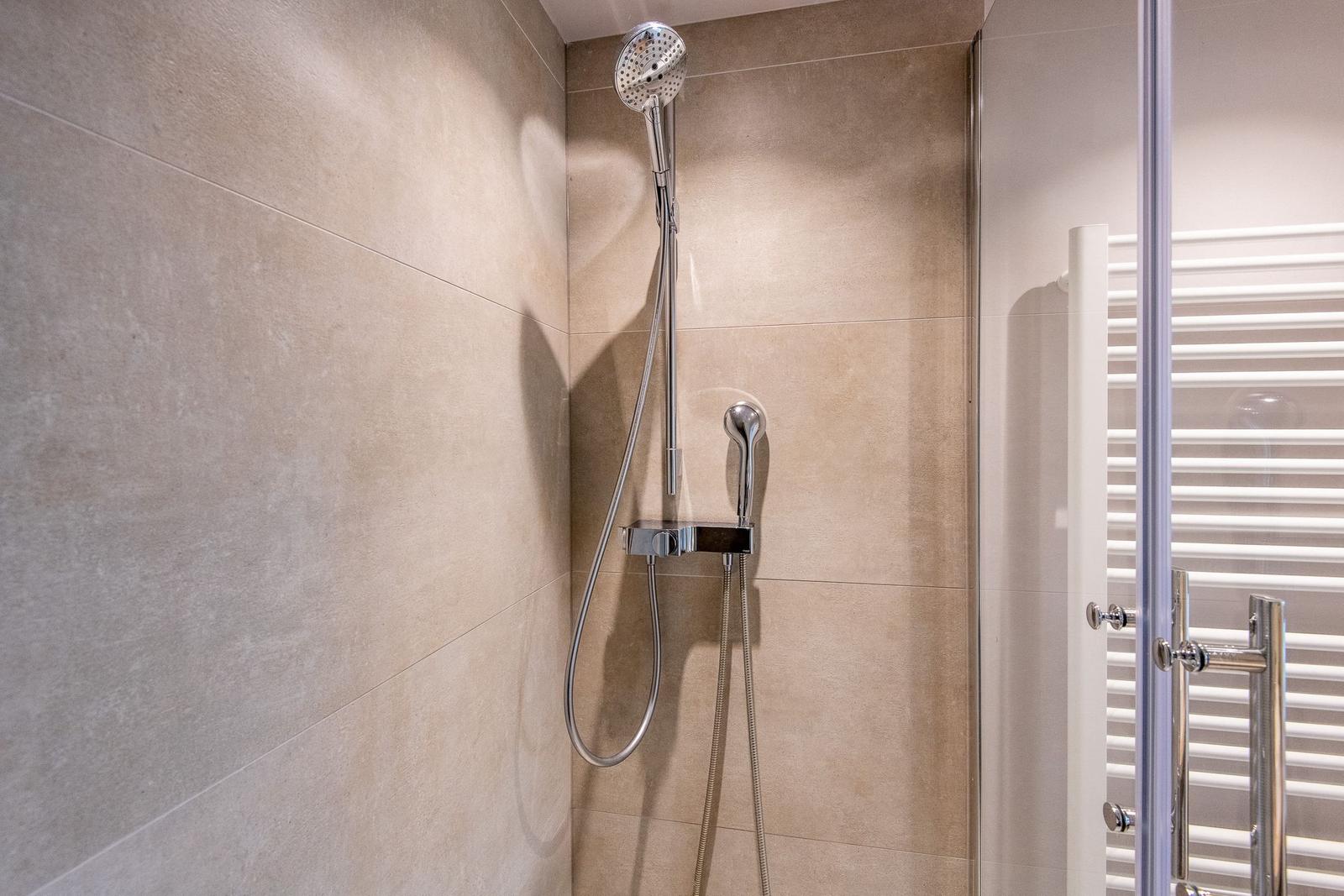 Dusche mit Handbrause und Duschwand aus hellen Fliesen.