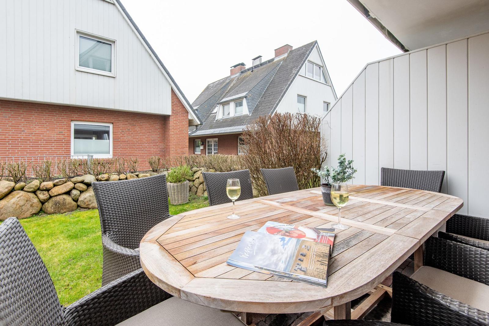 Terrasse mit Holztisch, Stühlen und Wein. Hintergrund: Haus und Garten.