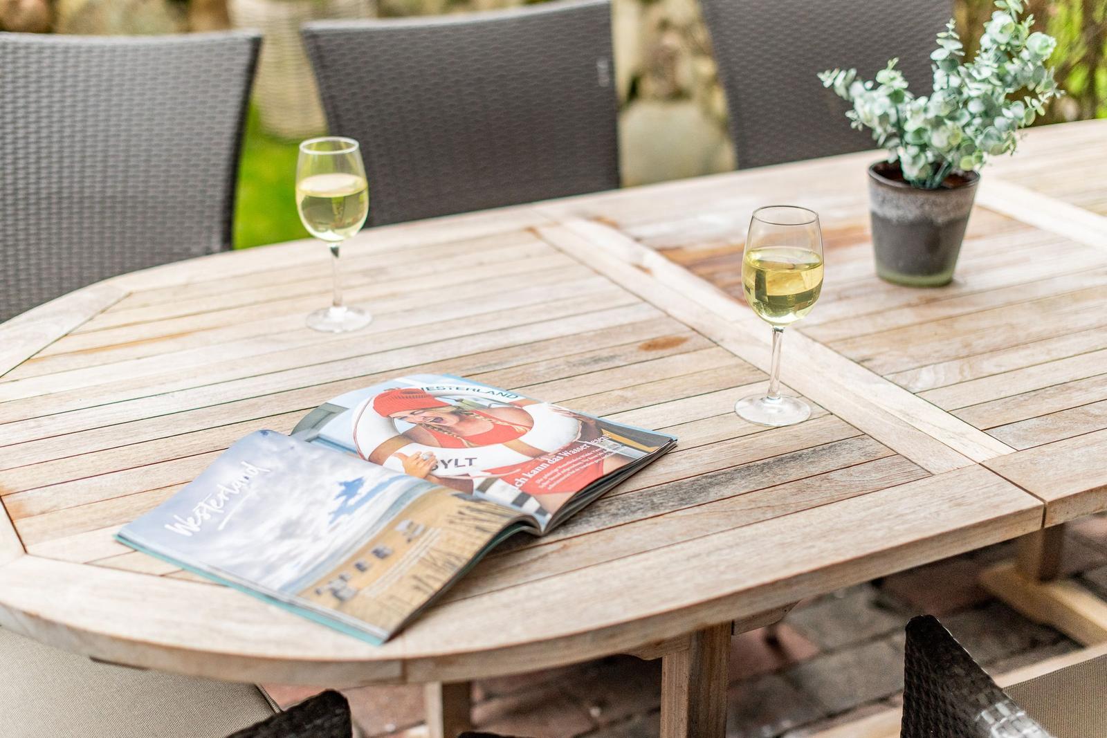 Holztisch mit zwei Weingläsern, Zeitschrift und Pflanze im Freien.