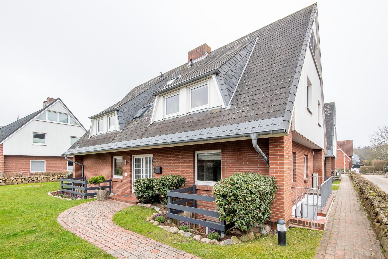 Ein zweistöckiges Haus mit rotem Backstein und grauem Dach, umgeben von Grünflächen und einem gepflasterten Weg.