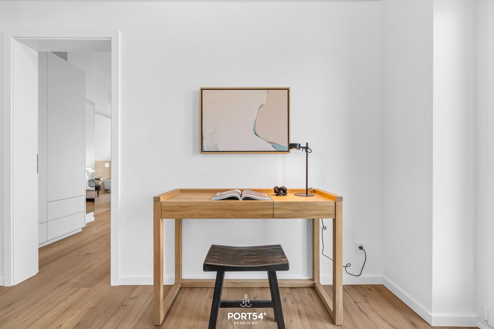 Schreibtisch mit Stuhl, Lampe und Bild an der Wand. Durchgang zu Schlafzimmer.