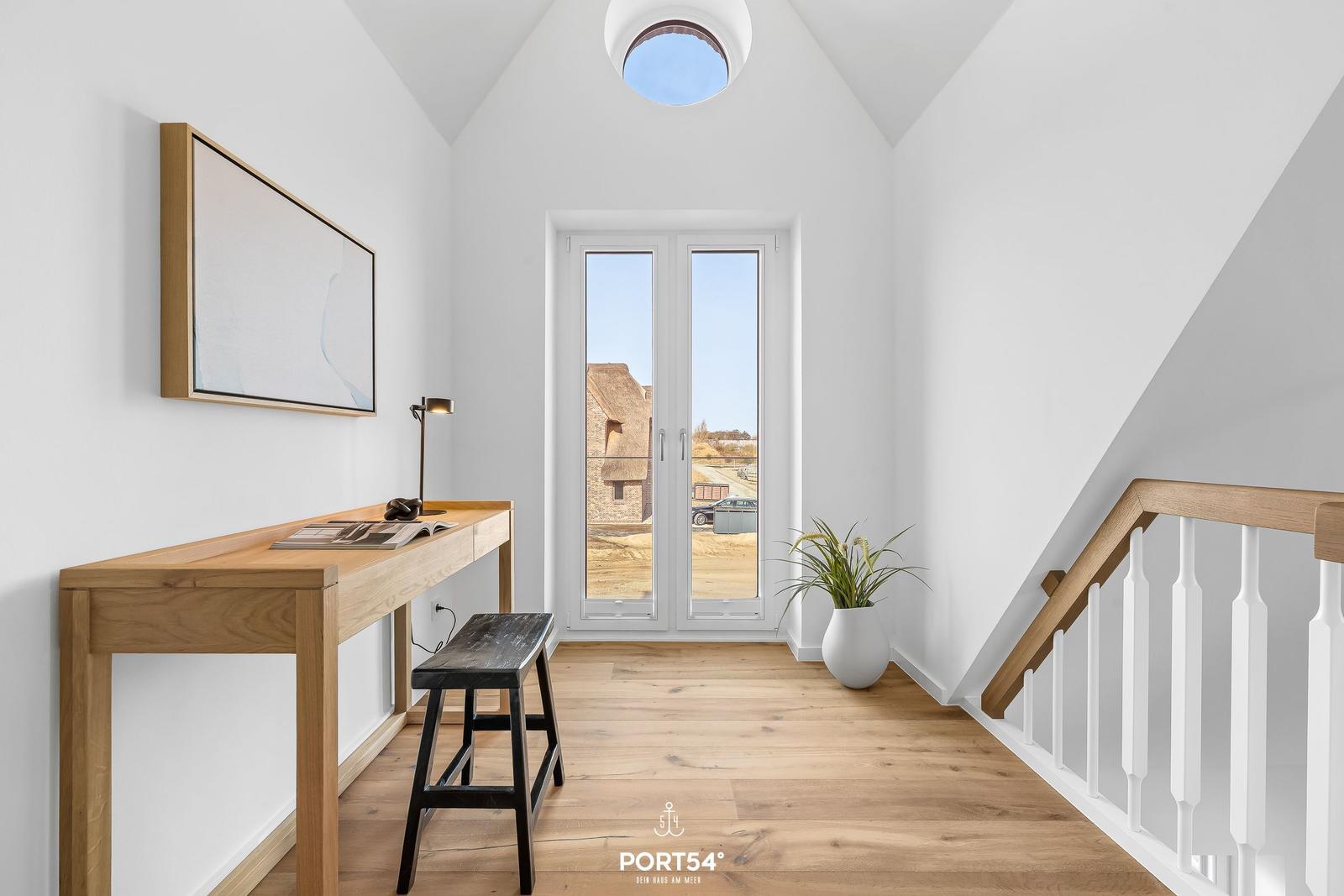 Helle Eingangshalle mit Schreibtisch, Pflanze und Treppe. Durch Fenster Blick auf Dachstuhl.