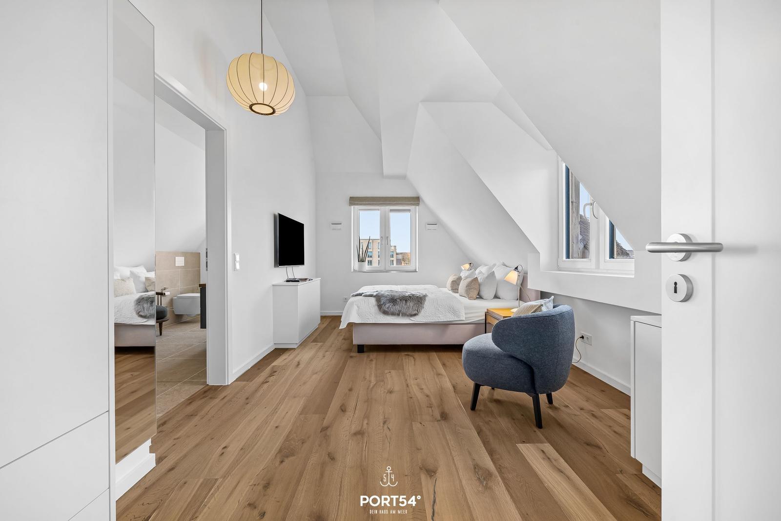 Schlafzimmer mit Dachschrägen, Bett, Stuhl und TV. Holzfußboden und Fenster.