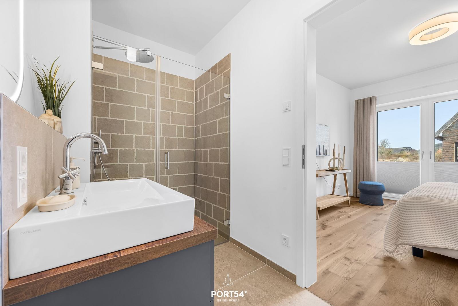 Badezimmer mit Dusche und Waschbecken. Nebenraum mit Bett und Fenster.