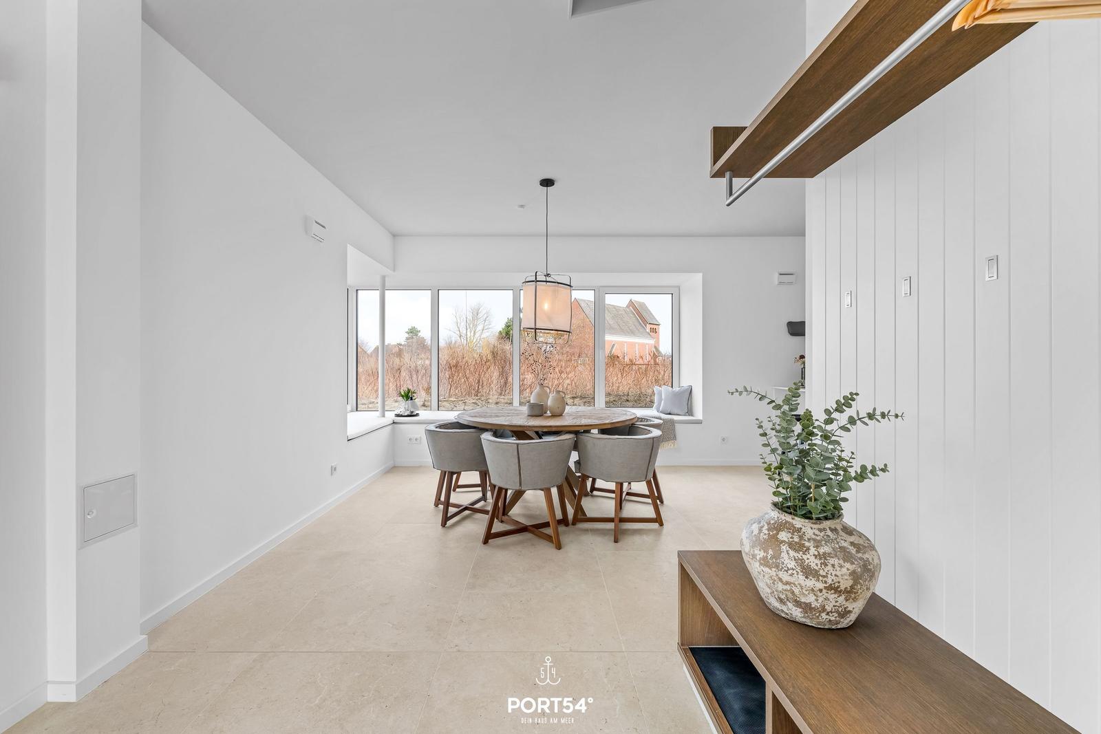 Dining room with round table, four chairs, and large window. Wooden shelf with plant on right.