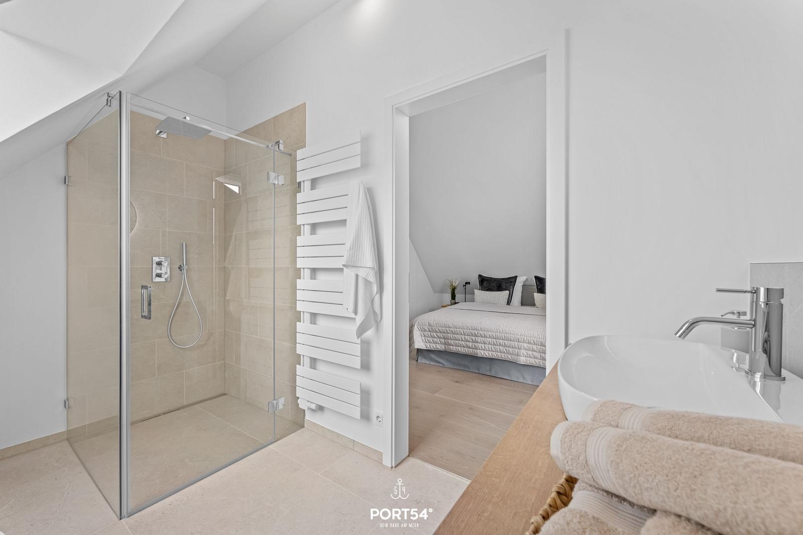 Badezimmer mit Dusche und Waschbecken. Schlafzimmer mit Bett im Hintergrund.