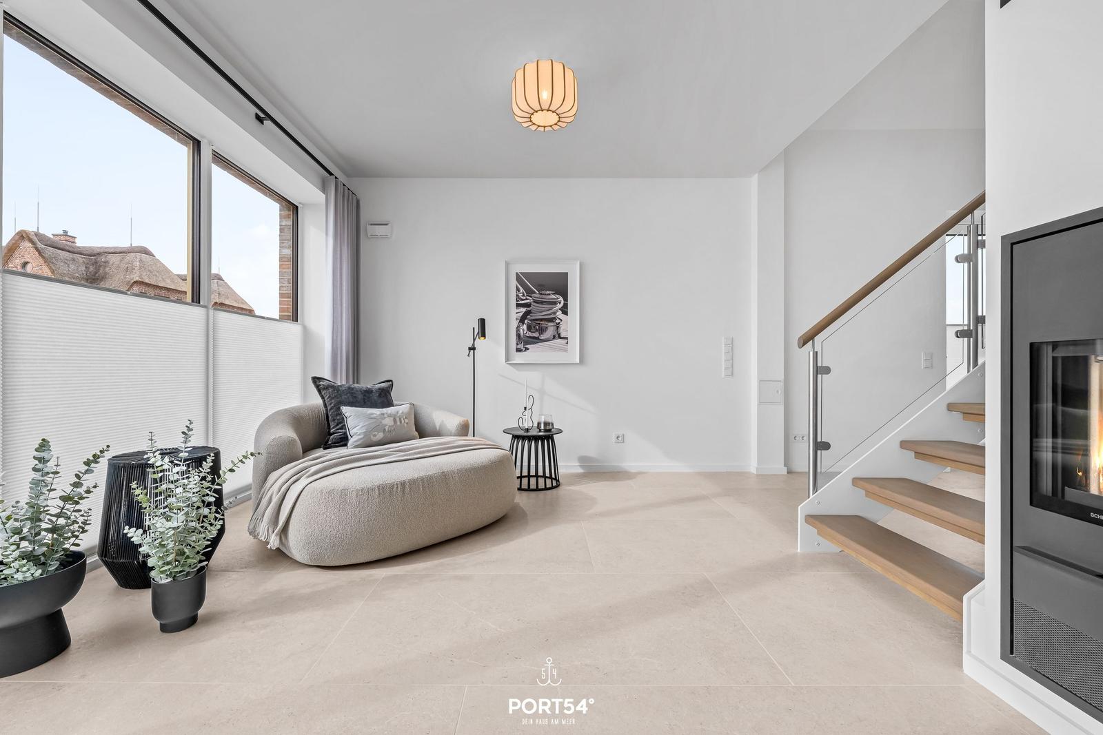 Wohnraum mit Sessel, Kamin und Treppe. Große Fenster und Pflanzen.