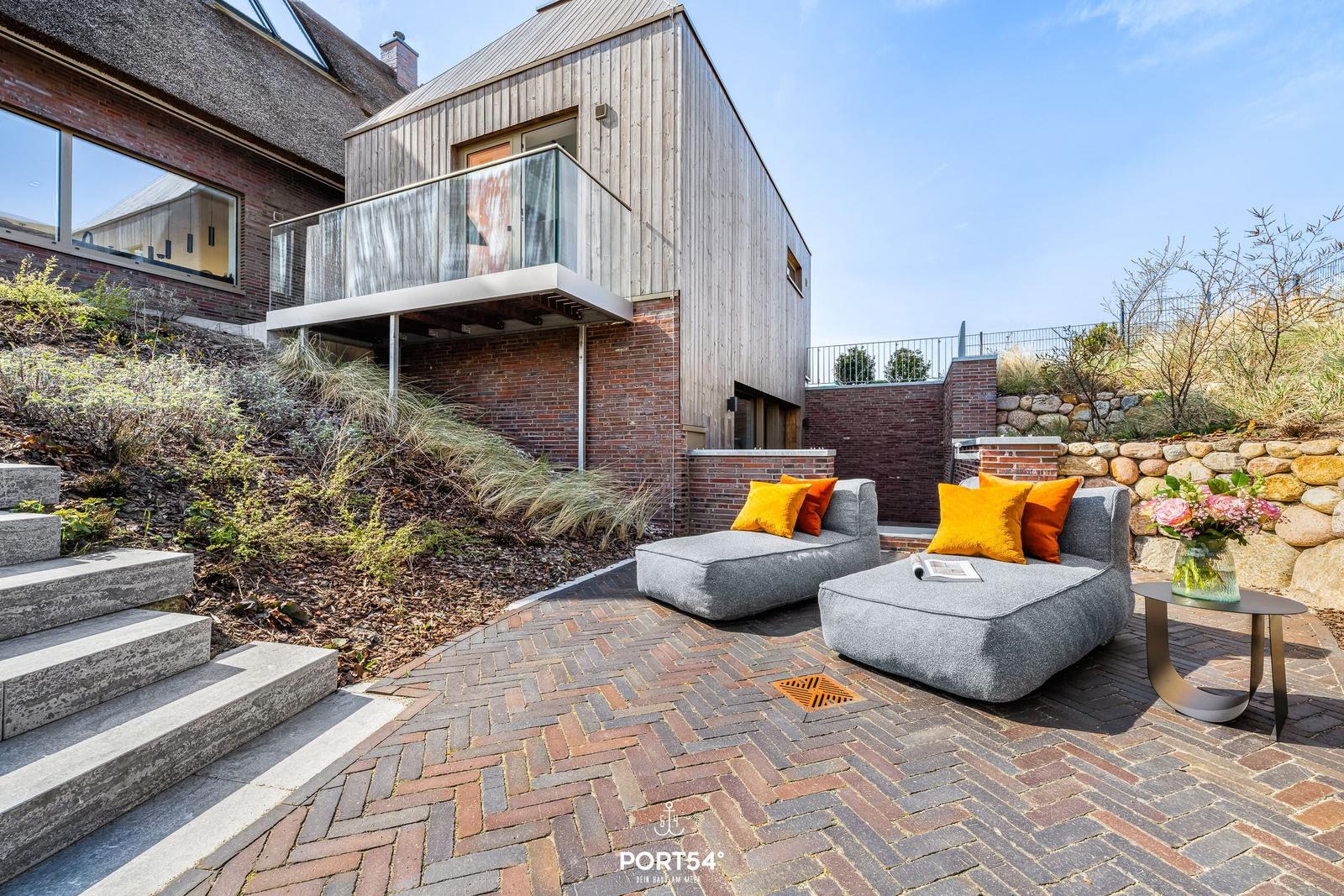Terrace with gray lounge cushions and orange pillows. Background: modern house with wooden facade.