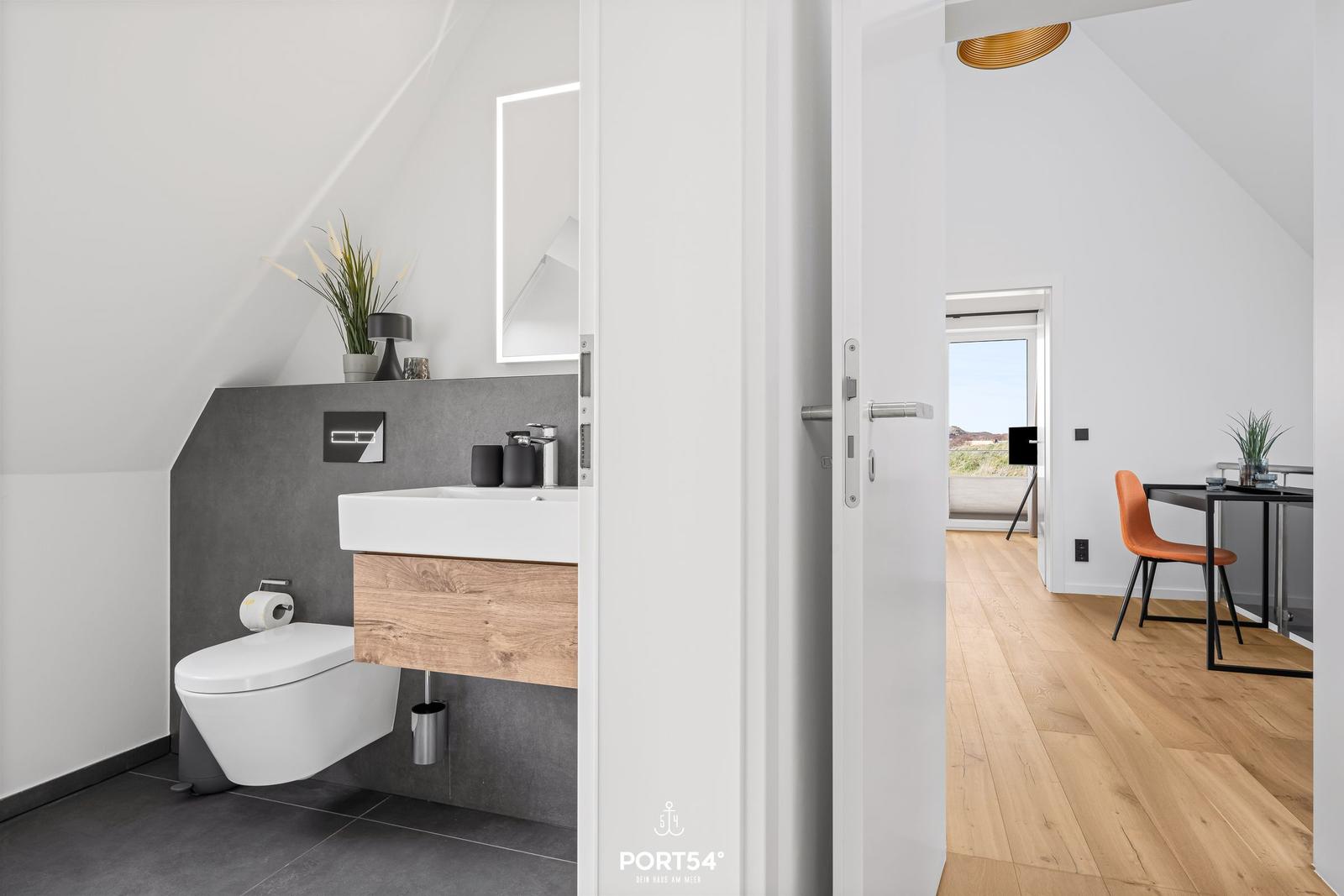 Bathroom with toilet and sink. View into living area with wooden floor and orange chair.