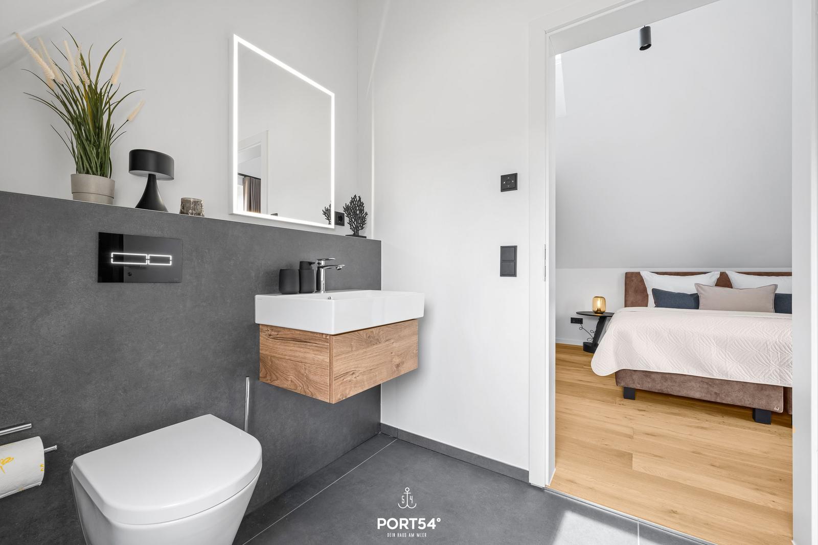 Bathroom with sink, mirror, and toilet. Bedroom with bed and wooden floor visible.