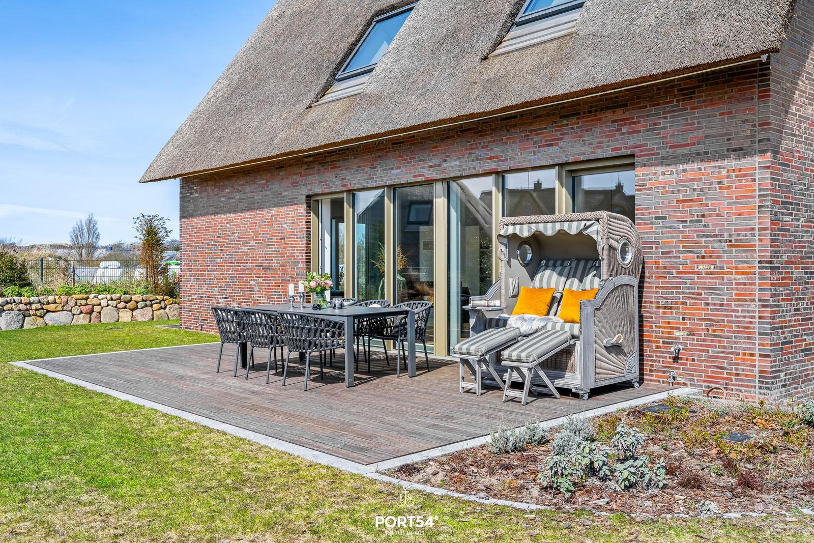 Deck with dining table, chairs, and beach chair in front of a thatched roof house.