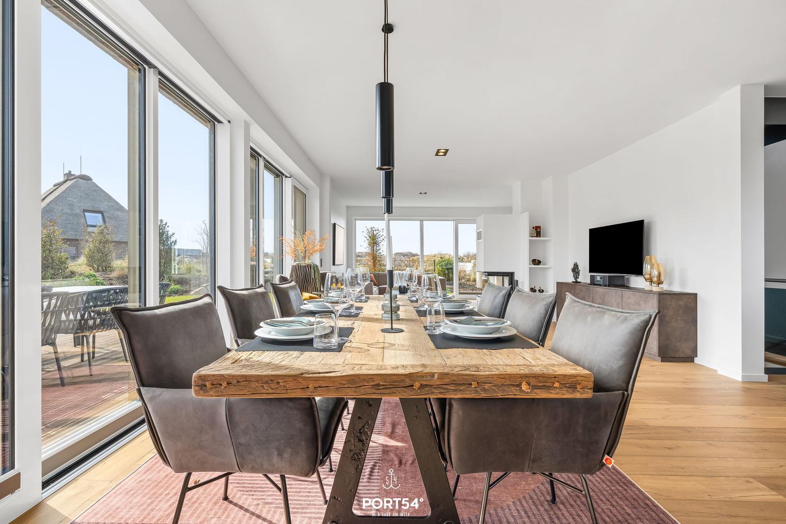 Dining area with long wooden table and chairs, large windows and TV.