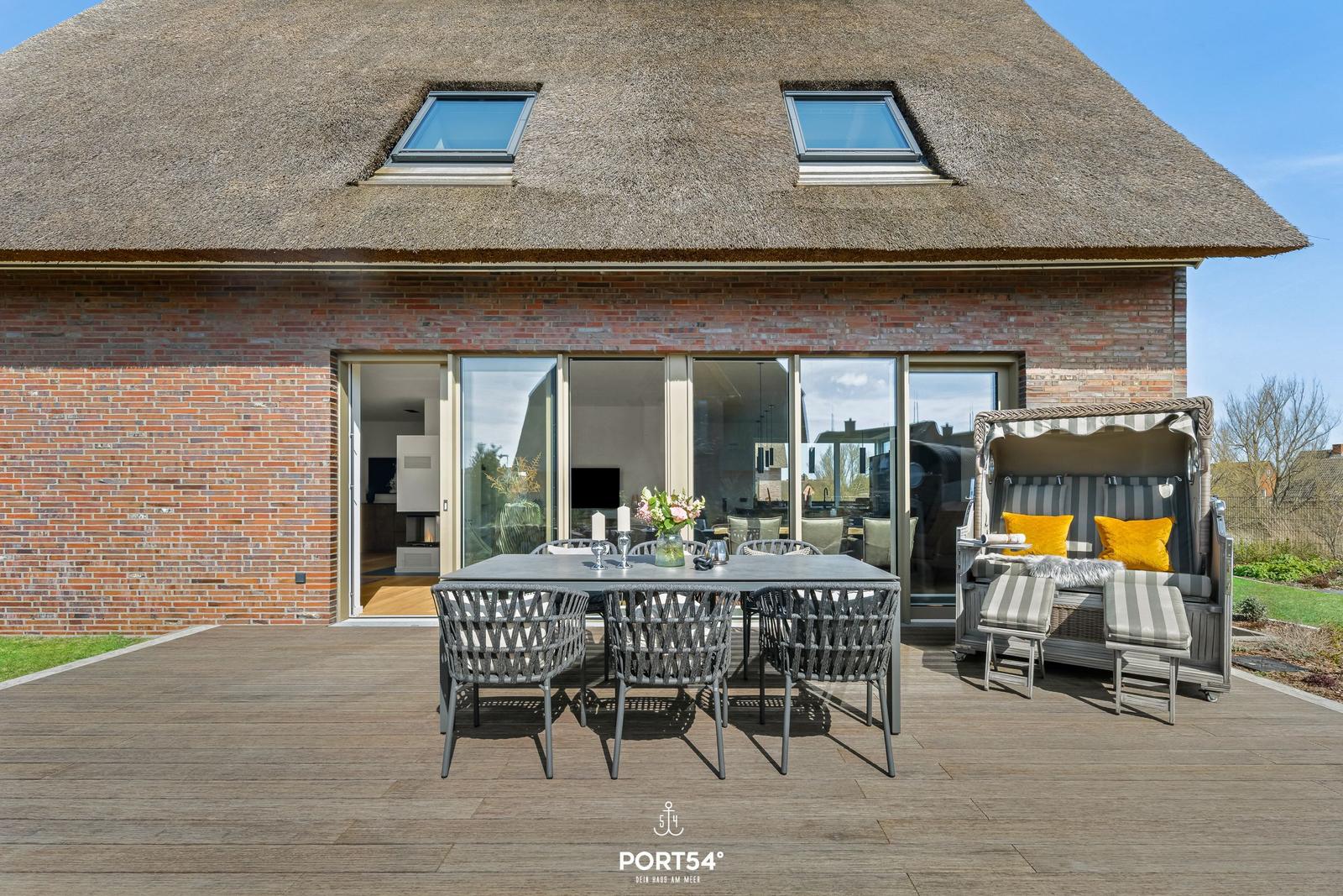 Terrace with dining area and beach chair in front of a house with thatched roof and glass facade.