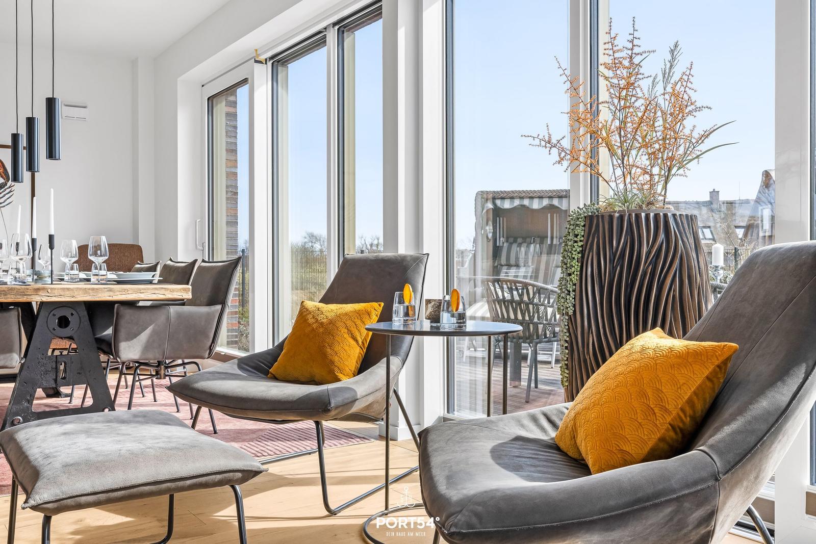 Two gray chairs with yellow cushions are by large windows. In the background, a dining area with table and chairs.