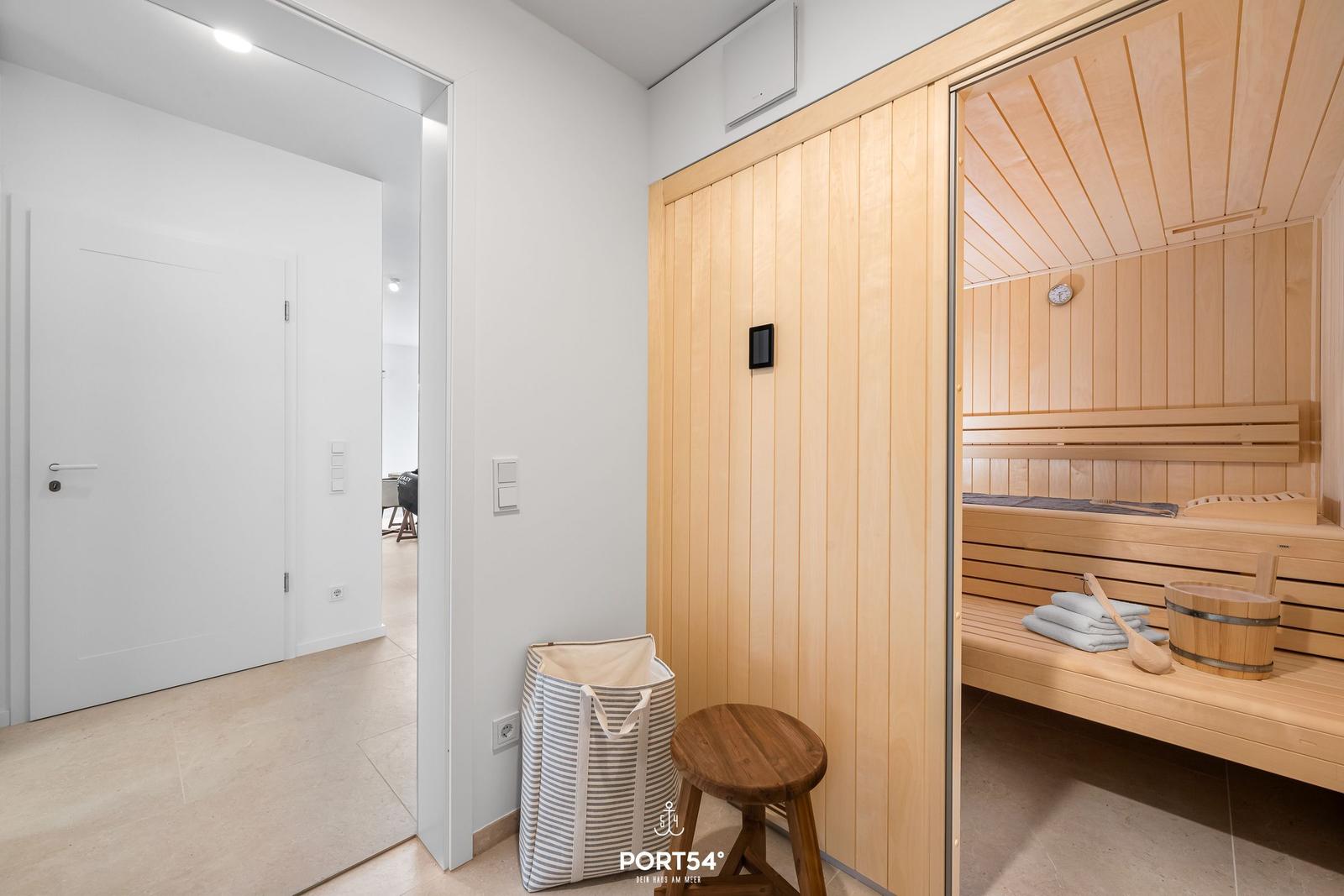 Sauna with wooden benches and bucket. Adjacent room with stool and laundry bag.