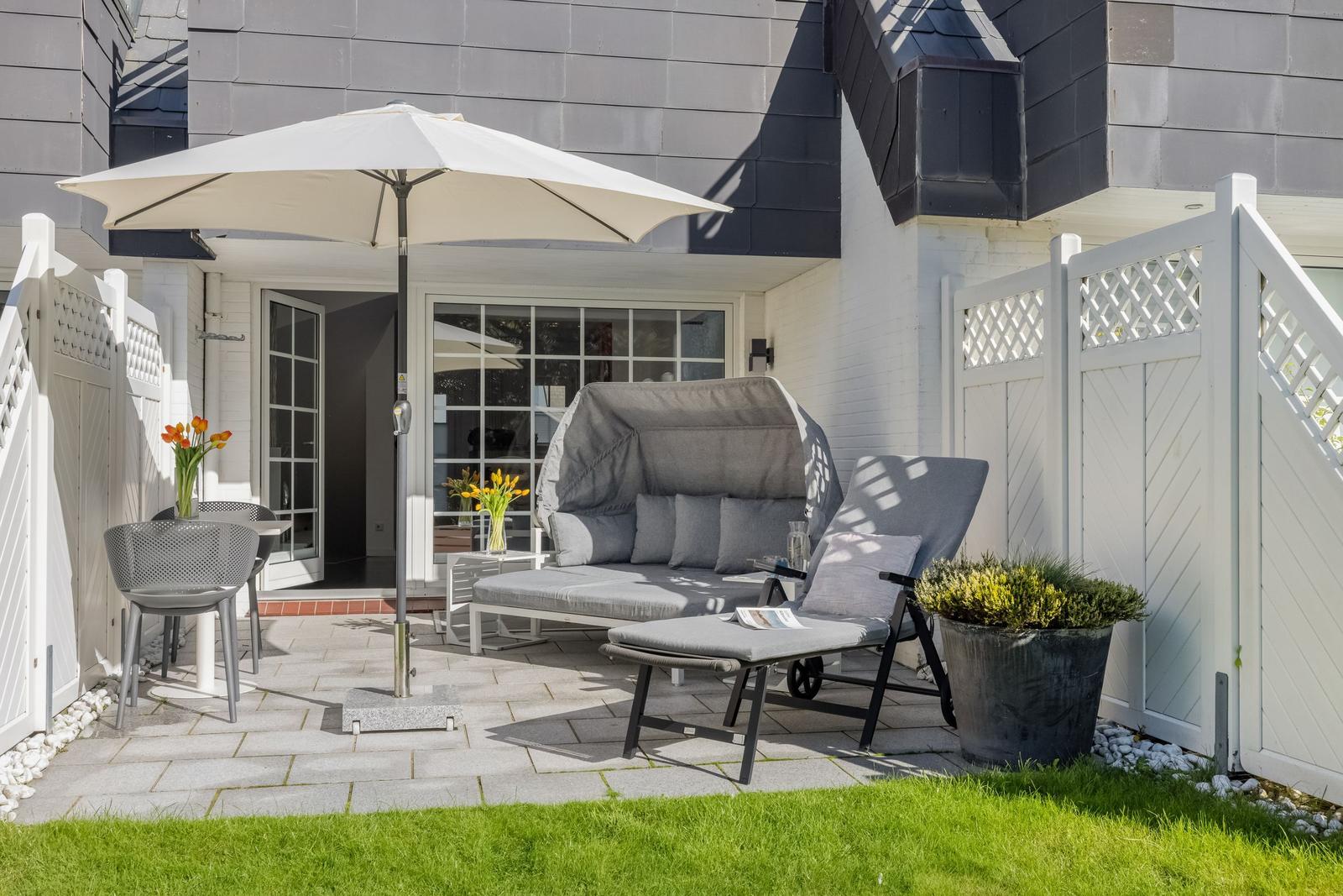 Terrace with seating, umbrella, and plants. Background: house with windows and white doors.
