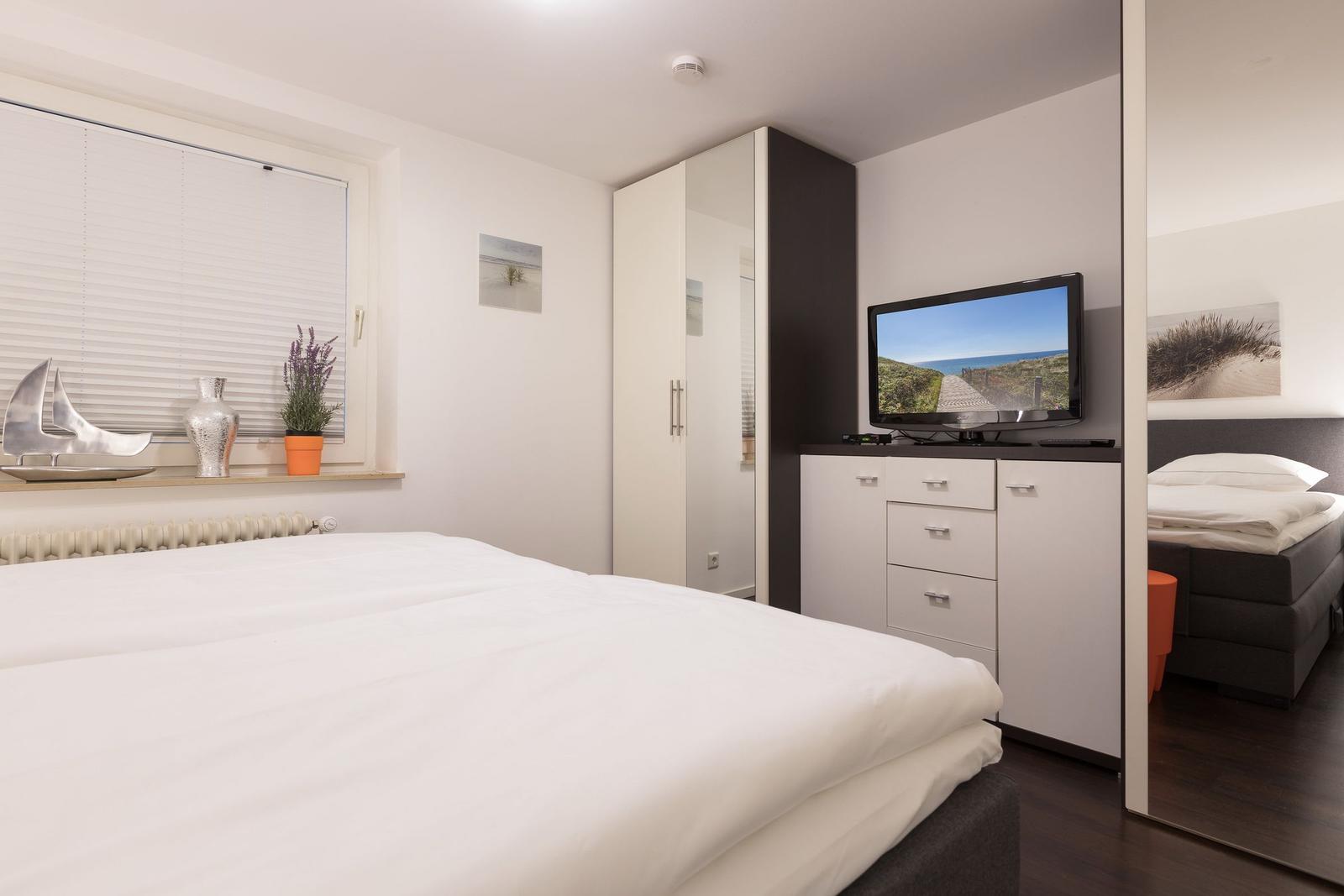 Bedroom with double bed, TV, and wardrobe. Window with blinds and plants.
