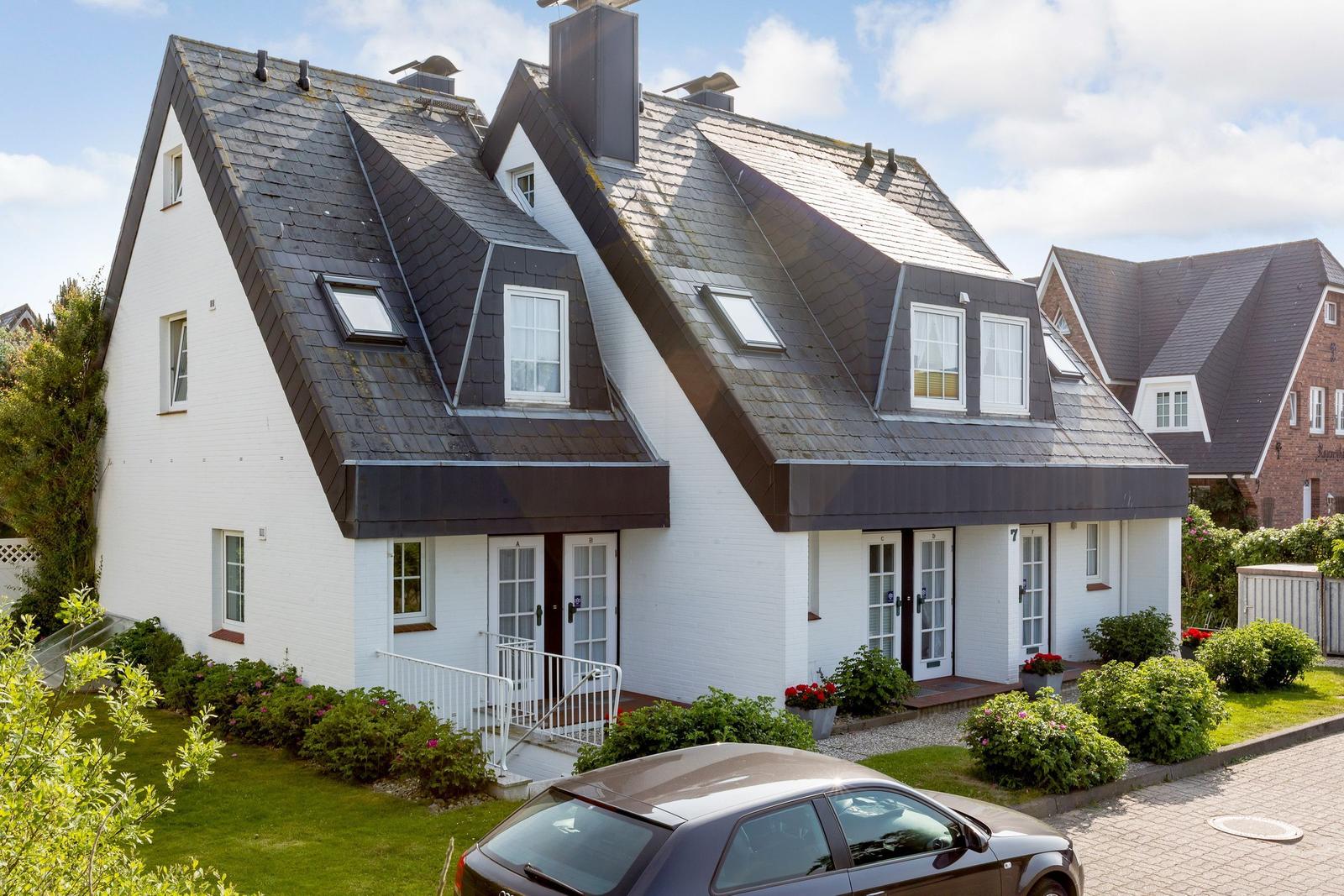 White villa with black roof and multiple windows. Front garden with flowerbeds and paved path.