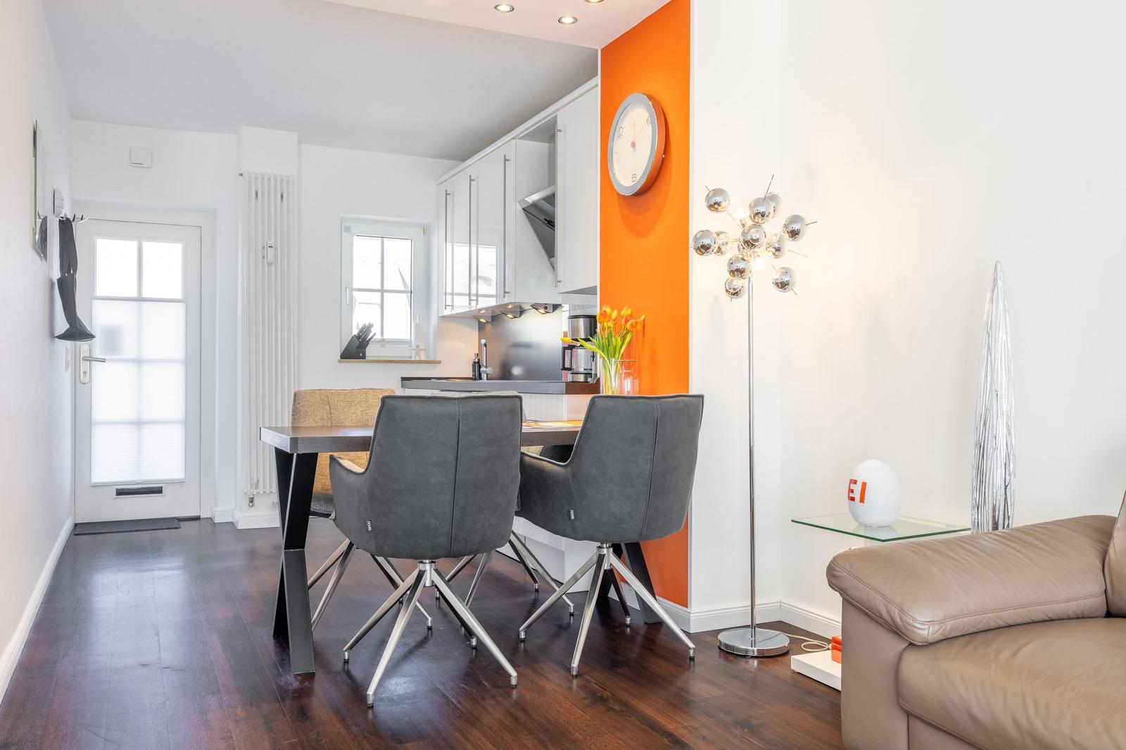 Dining area with table, chairs, and kitchen. Orange wall, lighting, and sofa visible.