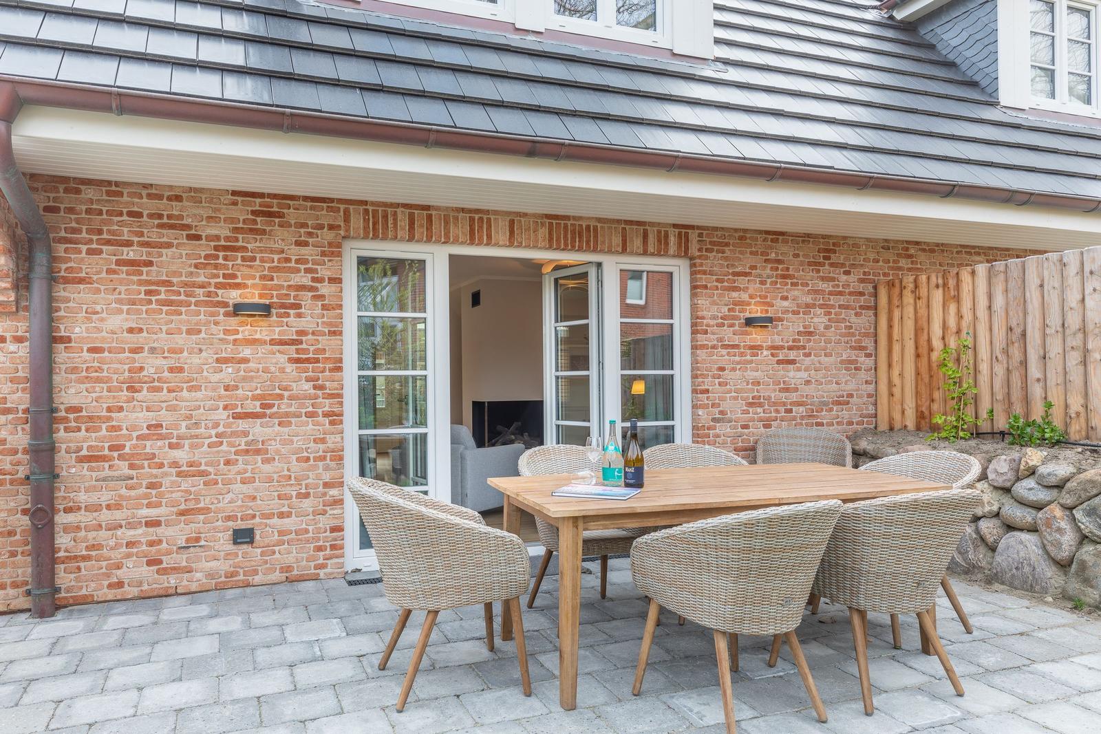 Terrace with wooden table and chairs in front of brick house with glass door.