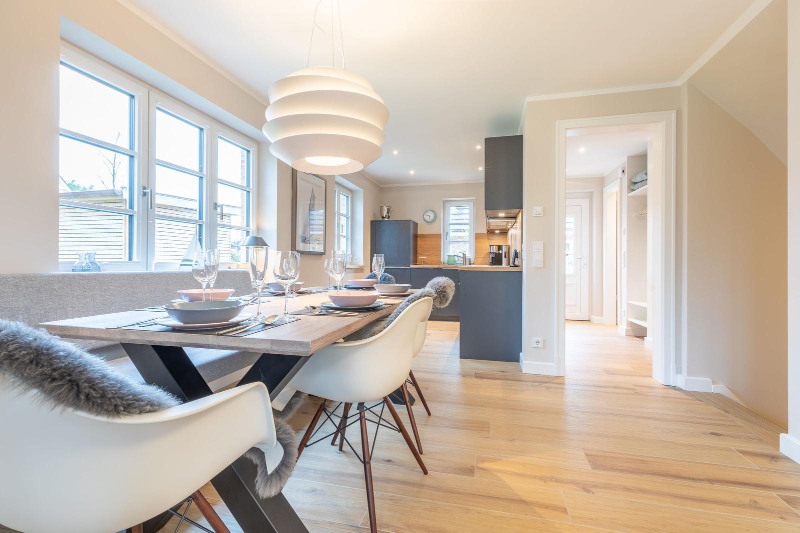 Dining area with table, chairs, and windows. Kitchen in background.