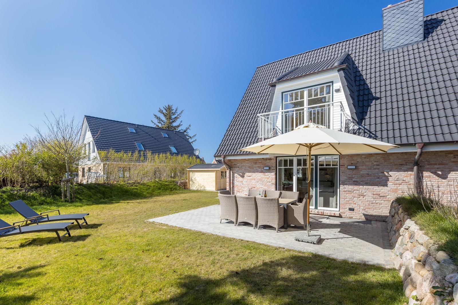 Haus mit Terrasse, Garten und Sonnenliegen unter blauem Himmel.