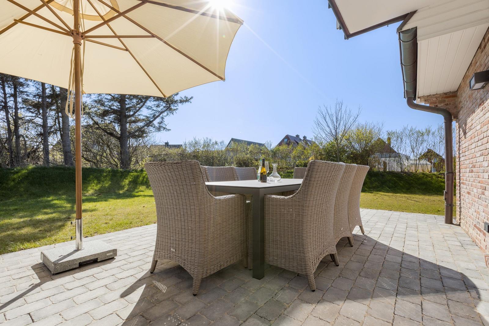 Terrasse mit Tisch, Stühlen und Sonnenschirm. Hintergrund: Gras, Bäume und Hauswand.