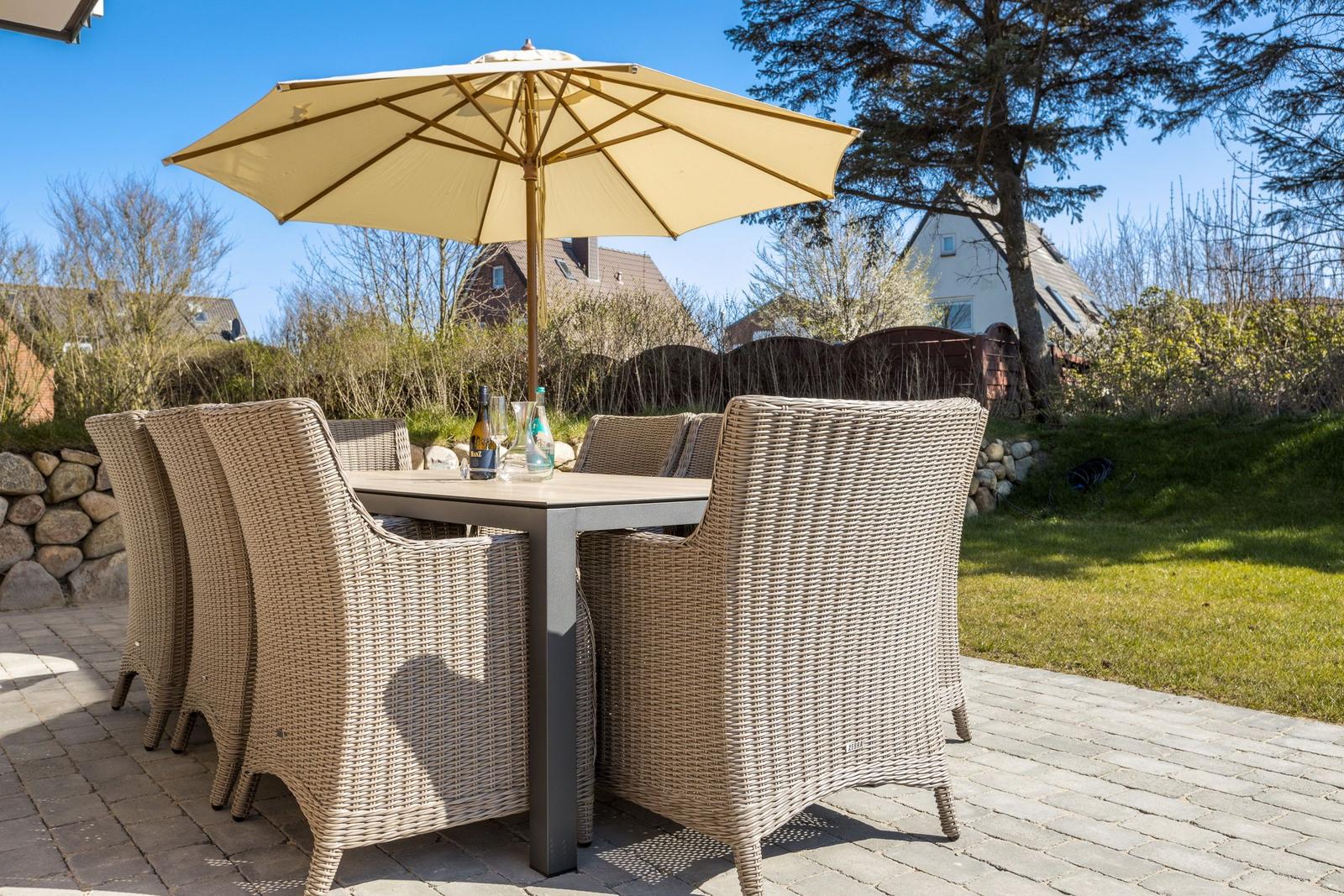 Terrasse mit Esstisch, sechs Stühlen und Sonnenschirm. Hintergrund: Grasfläche und Bäume.