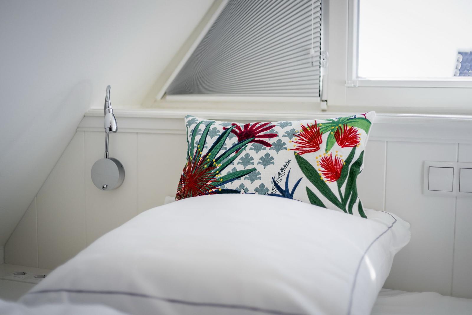 Schlafzimmer mit weißer Bettwäsche und buntem Kissen unter Dachschrägen.
