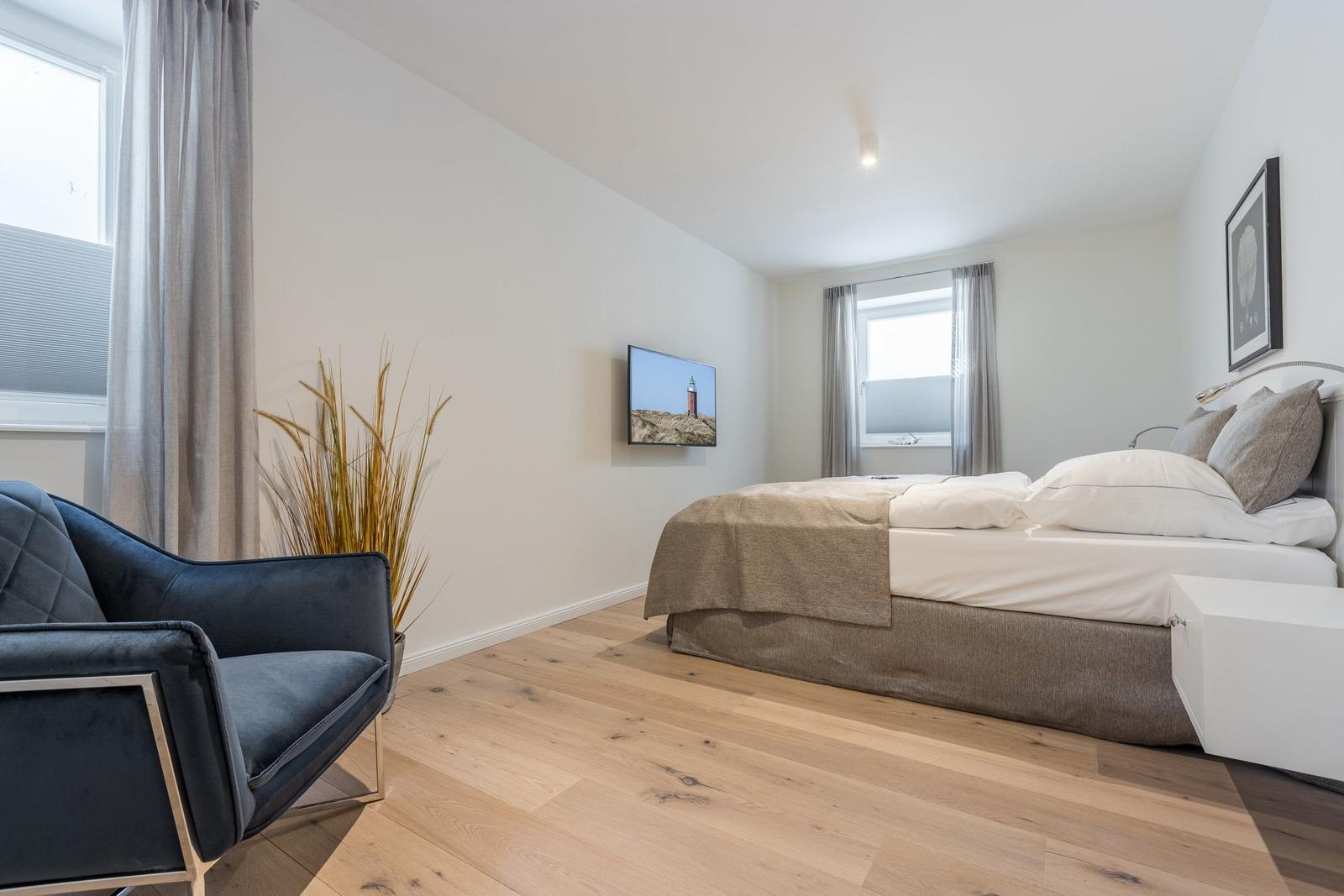 Schlafzimmer mit Bett, TV und Sessel. Holzboden und Fenster mit Vorhängen.
