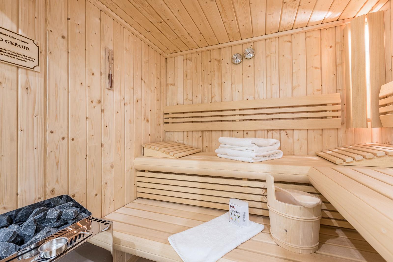 Holzsauna mit Bänken, Handtuch und Eimer. Ofen mit Steinen und Warnschild.