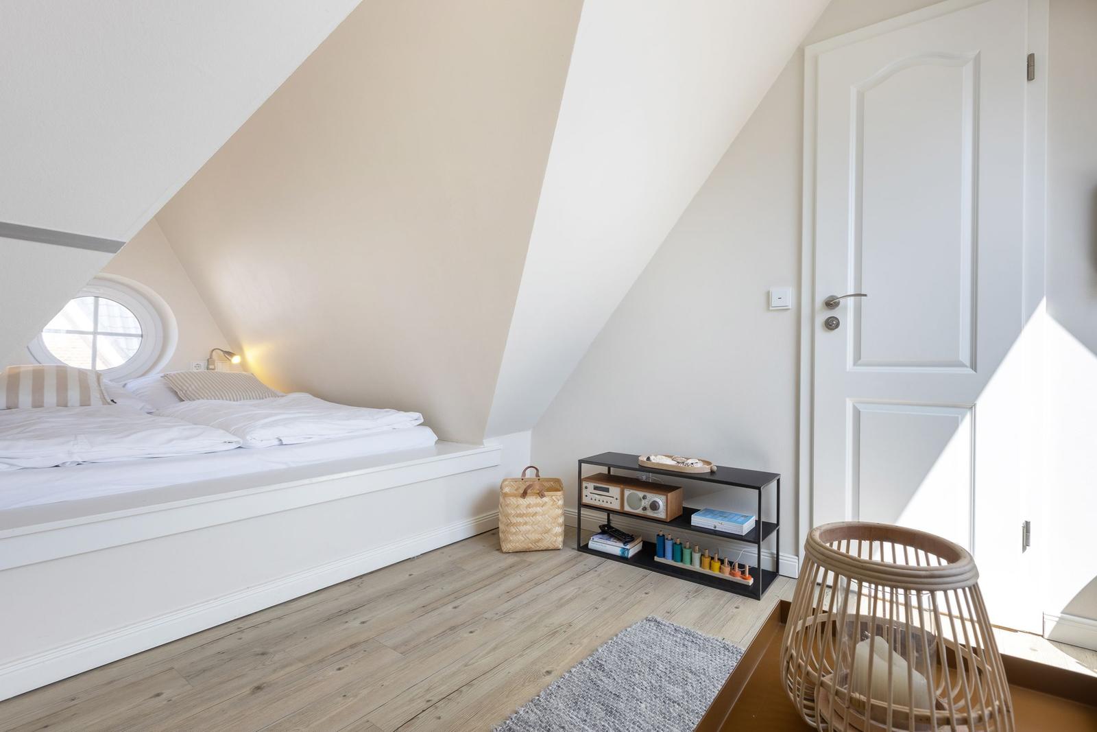 Bedroom with sloped ceiling, round window, bed, and wicker chair.