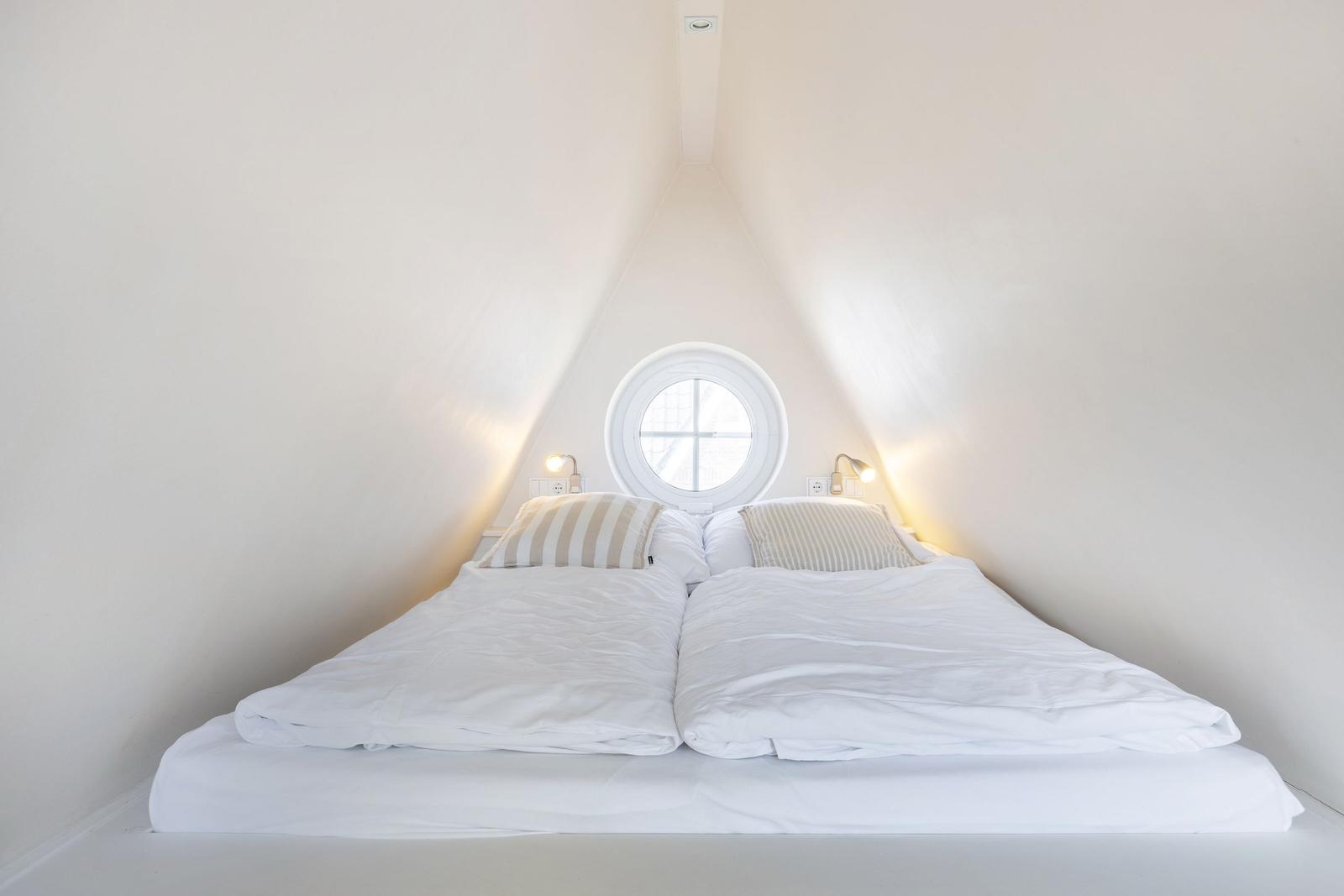 Double bed under sloped ceilings with round window and bedside lamps