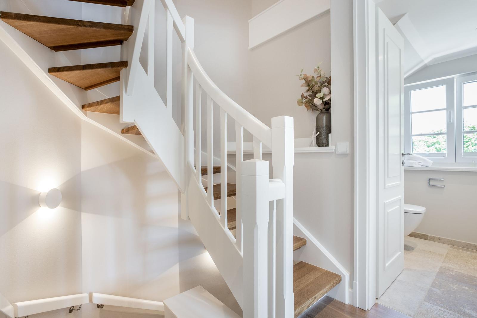Treppenhaus mit Holzstufen und weißer Balustrade. Neben dem Flur ist ein Badezimmer sichtbar.