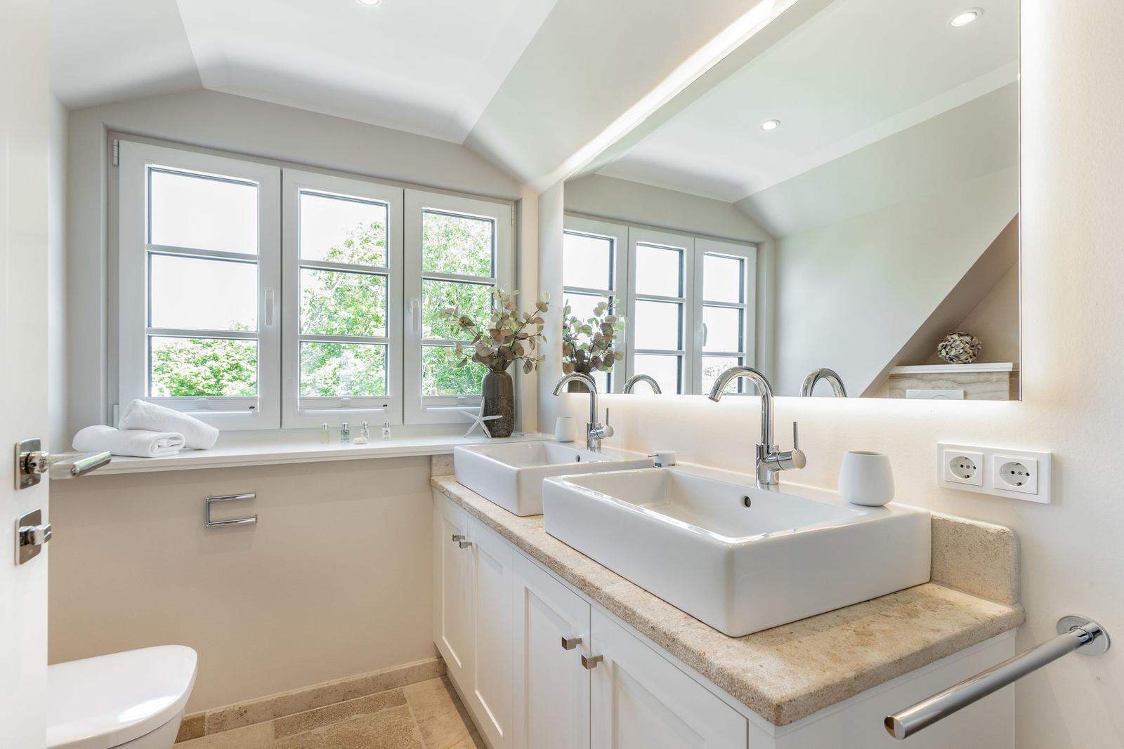 Badezimmer mit zwei Waschbecken, Spiegel und Fenster mit Blick auf Grün.
