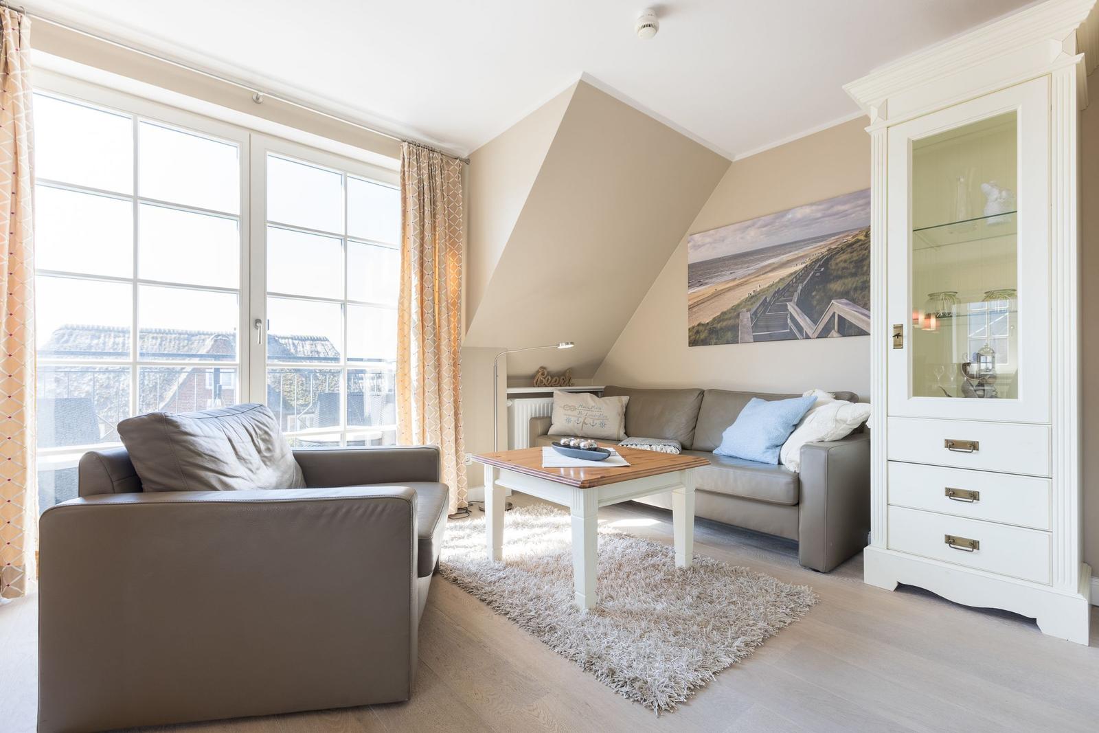 Living room with sofa, armchair, and table. Large windows and white cabinet.