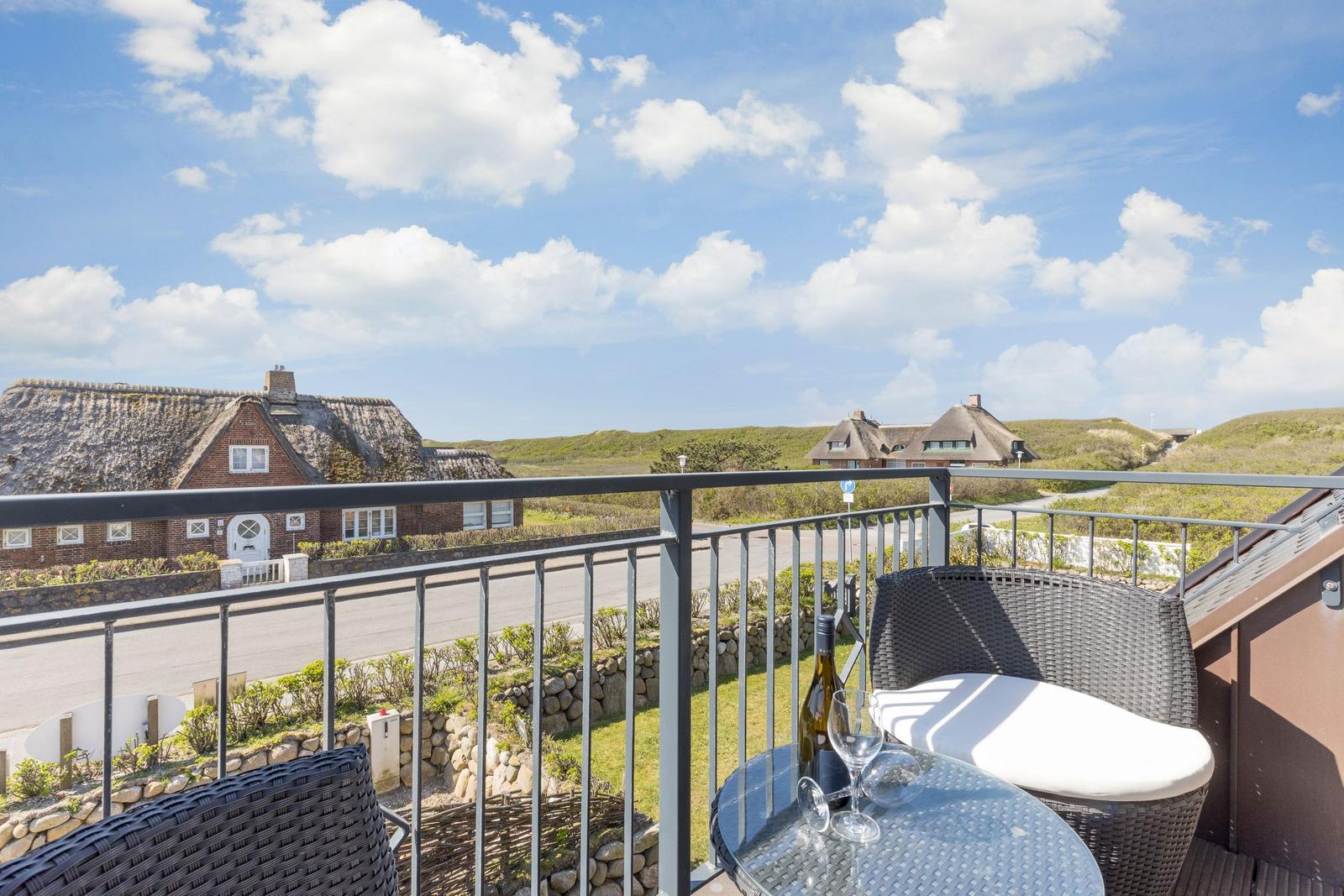 Balcony with table and chairs overlooking thatched-roof houses and green dunes.