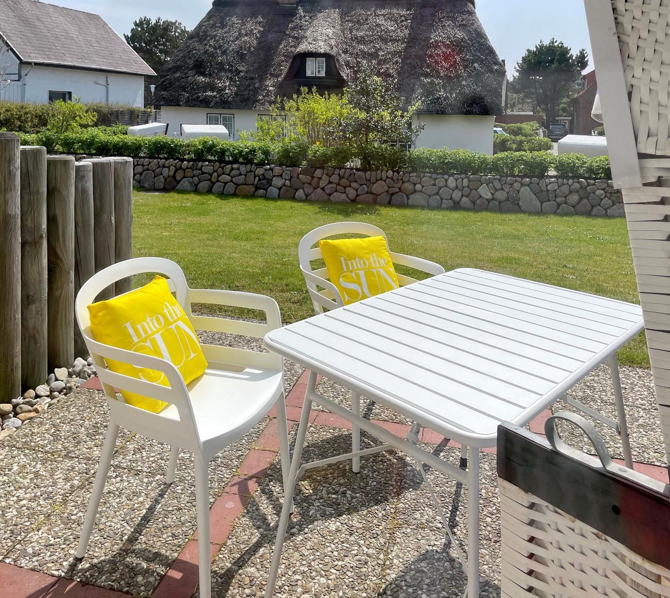 White table and two chairs with yellow cushions on terrace. Background: lawn and thatched house.