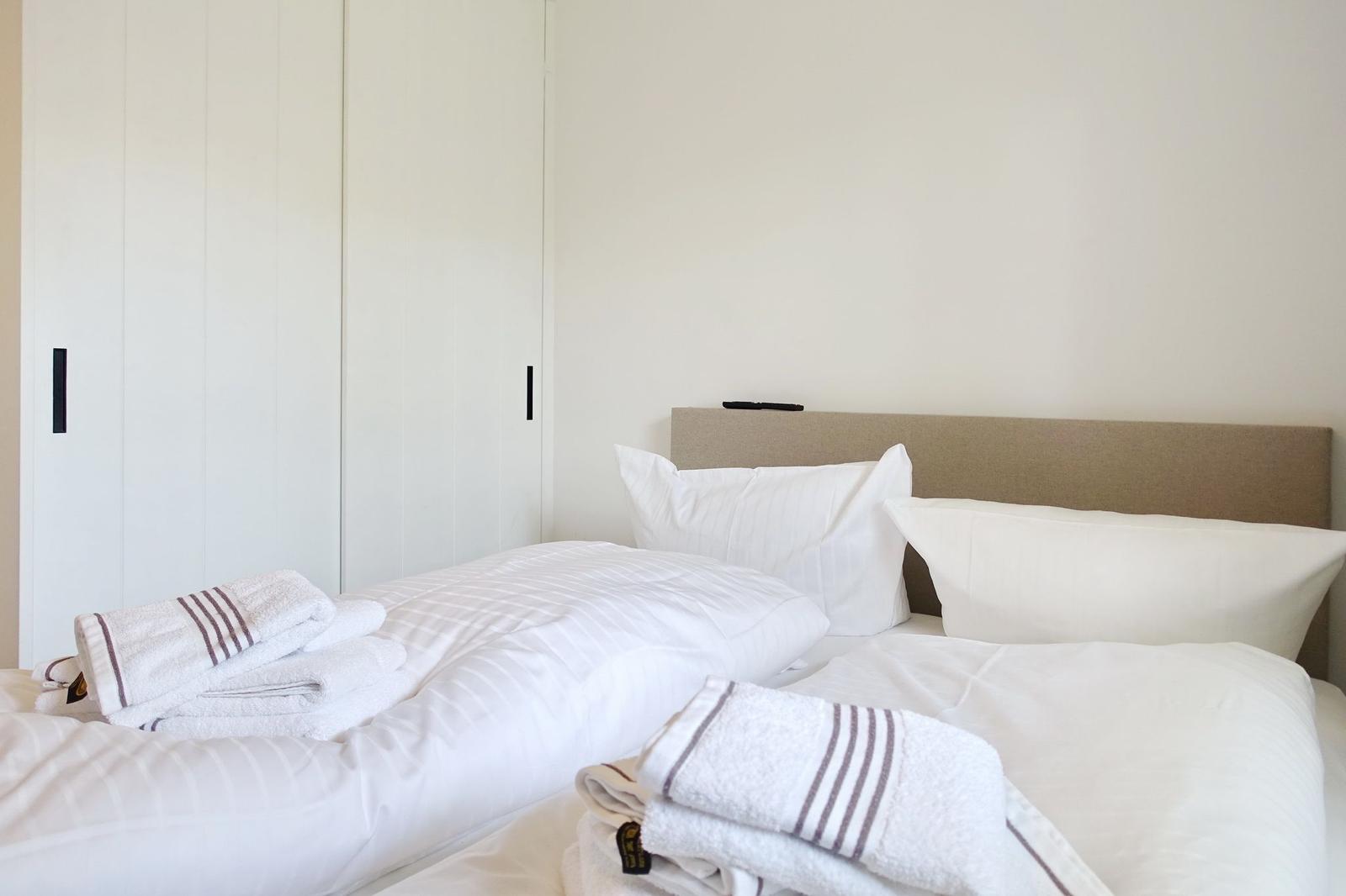 Bedroom with bed, pillows, and folded towels. A wardrobe stands behind the bed.