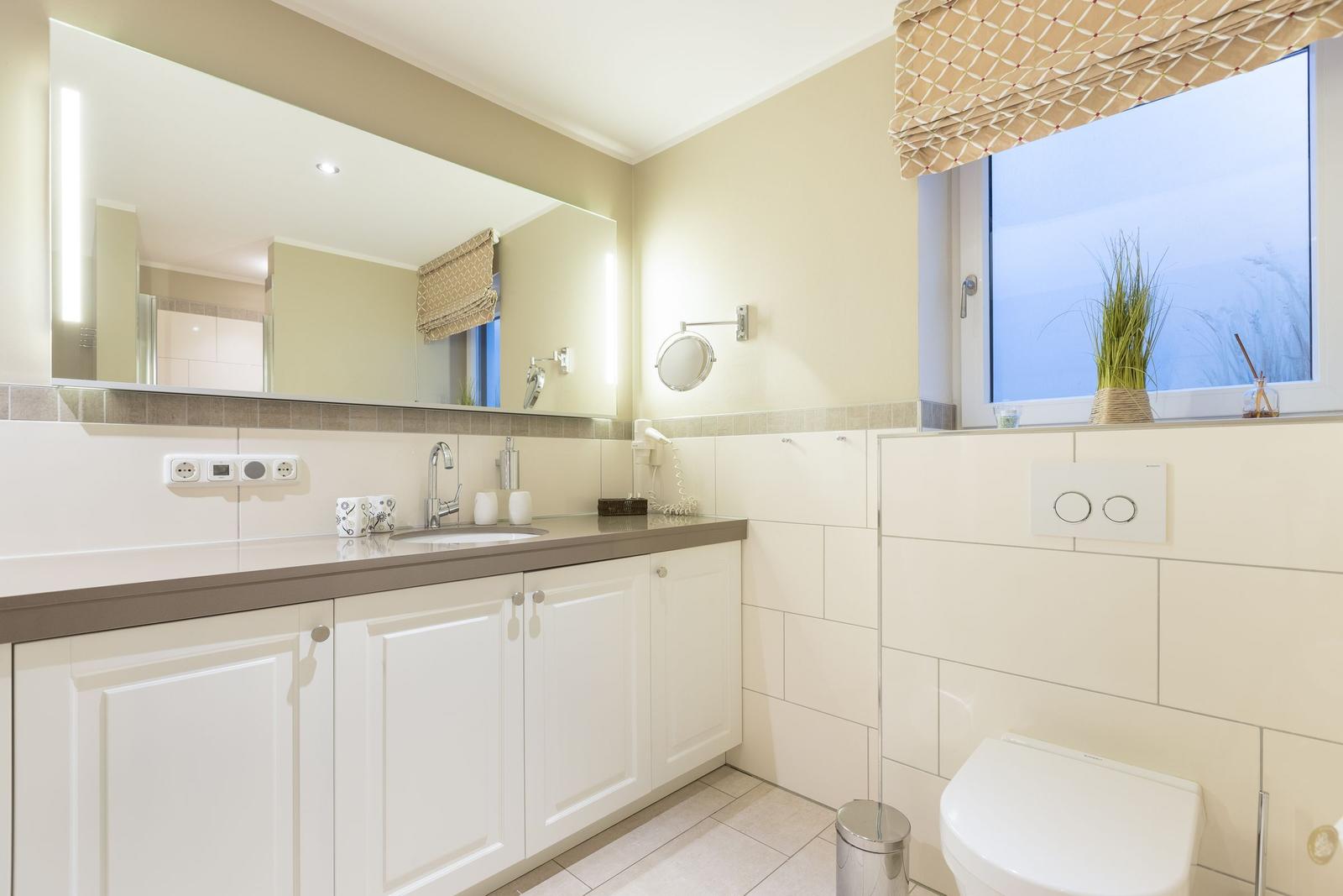 Badezimmer mit Waschbecken, Spiegel und Toilette. Fenster mit Pflanze.