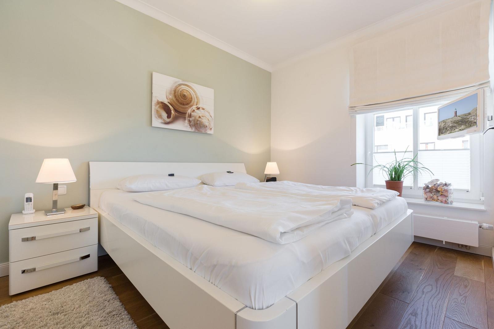 Schlafzimmer mit weißem Bett, Nachttisch und Fenster mit Pflanze.