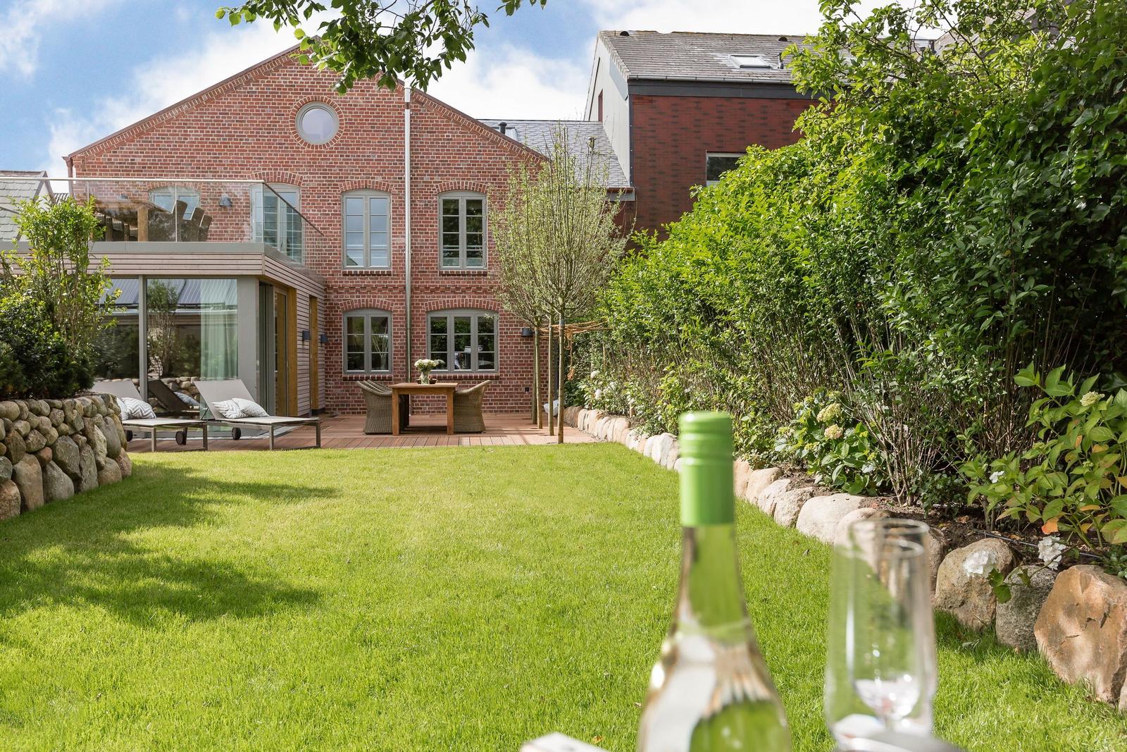 Red brick house with garden, terrace, and green lawn. Foreground: wine bottle and glass.