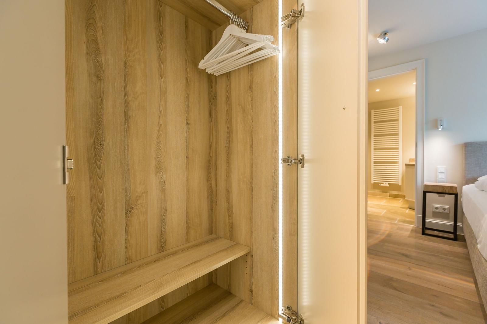 Closet with wooden walls and hangers. View into bedroom with bed and nightstand.